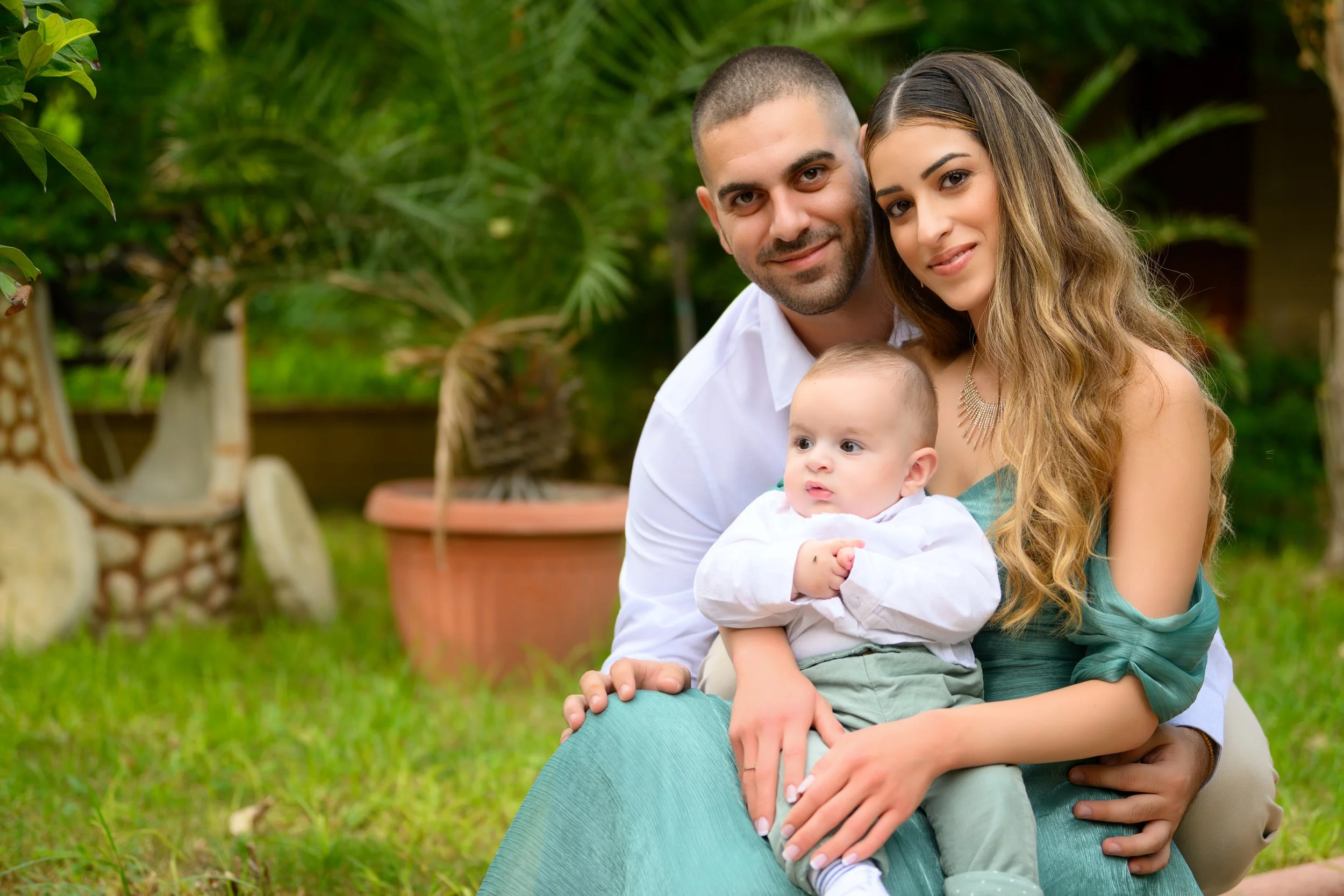 A family of three sitting outdoors in a garden with green plants, a mother, father, and a baby boy. The father is behind and to the left, the mother is on the right, and the baby is in front, held by both parents.