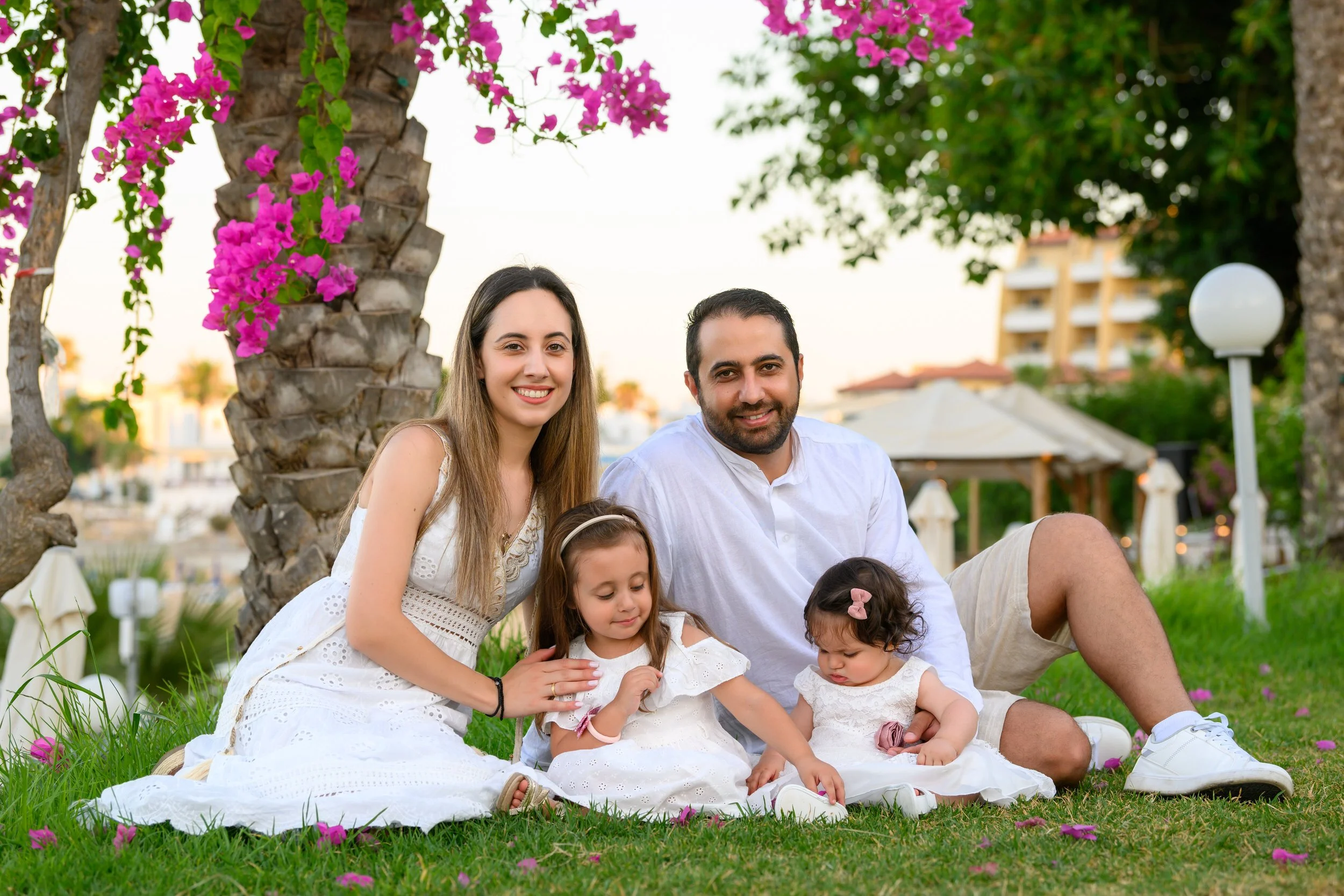 A family of four sitting on the grass beneath a palm tree with pink flowers, with two young girls and a smiling woman and man, outdoors during daylight.