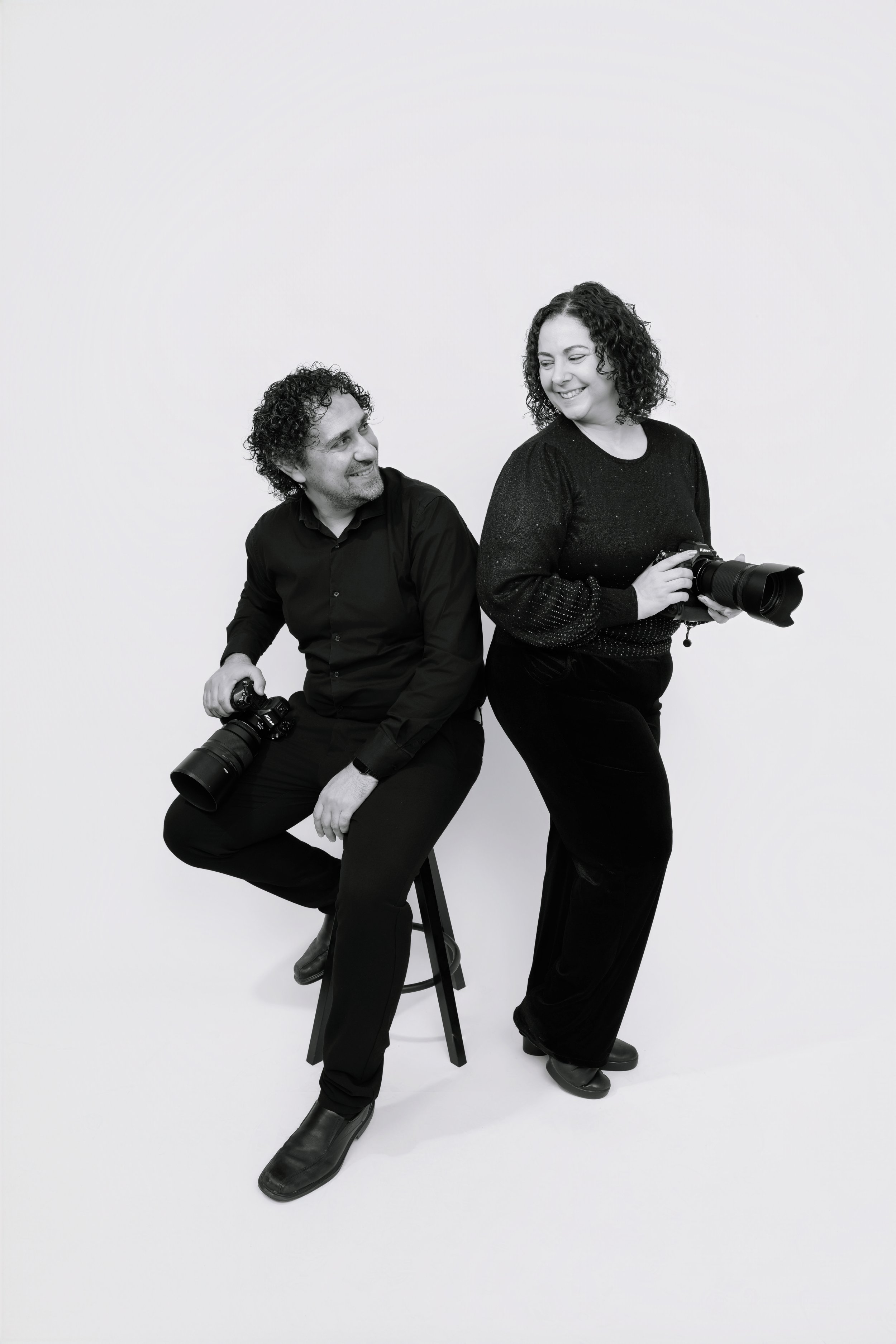Two people, a man and a woman, holding cameras, smiling and looking at each other against a plain white background. The man is sitting on a stool, and the woman is standing beside him.