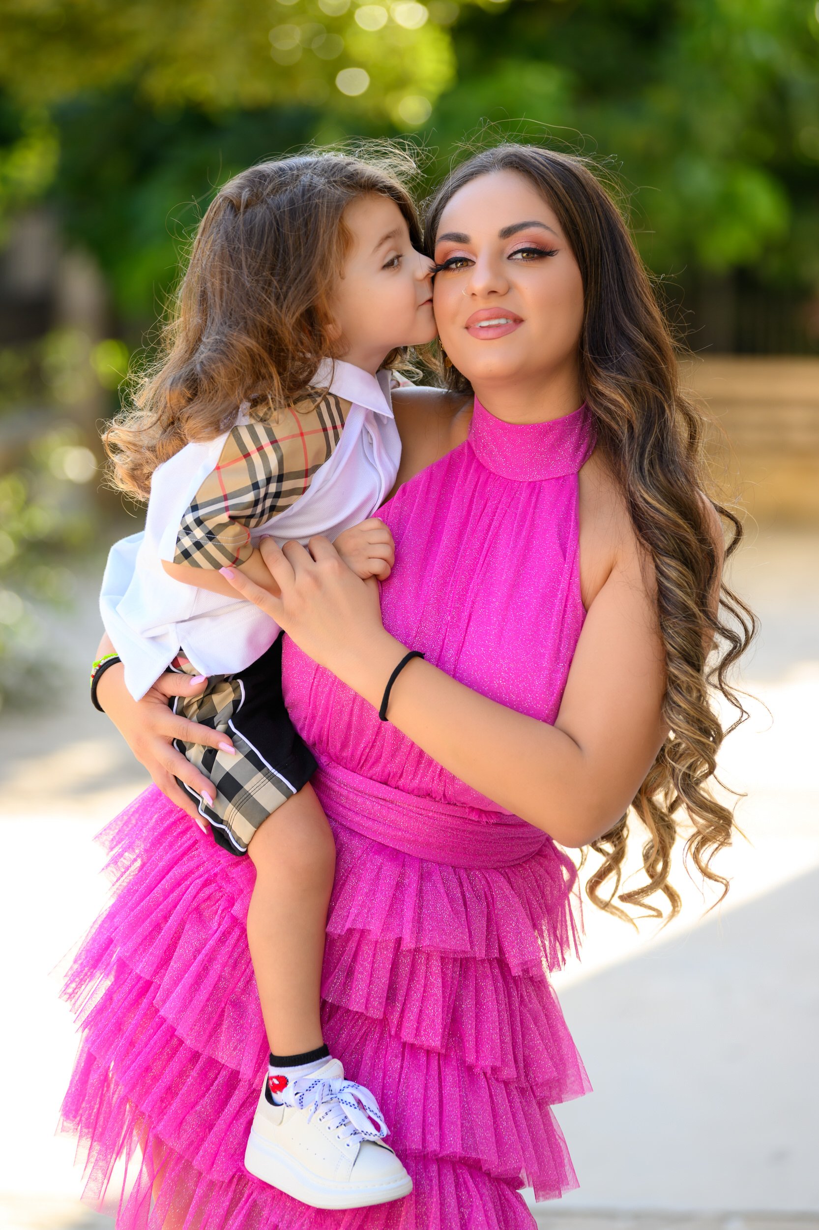 Young woman in a pink dress holding a young girl in a white shirt with plaid sleeves, outdoors with greenery.