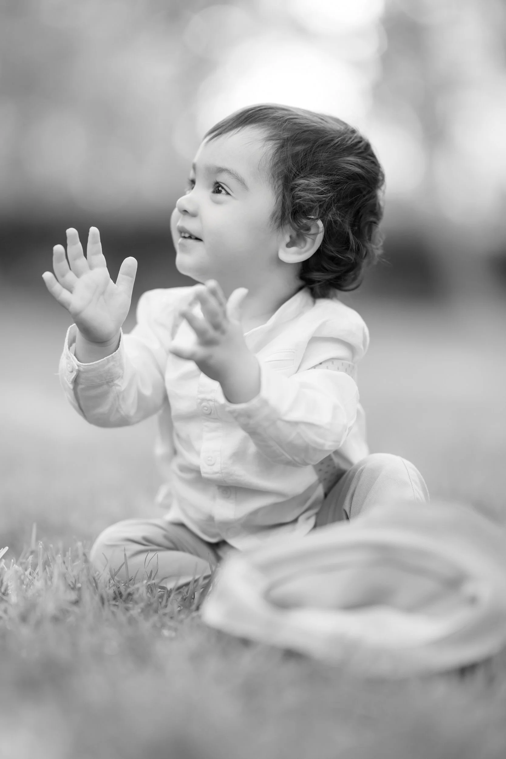 A young child with dark, curly hair sitting on grass, clapping and smiling outdoors, blurred natural background, black and white photograph.
