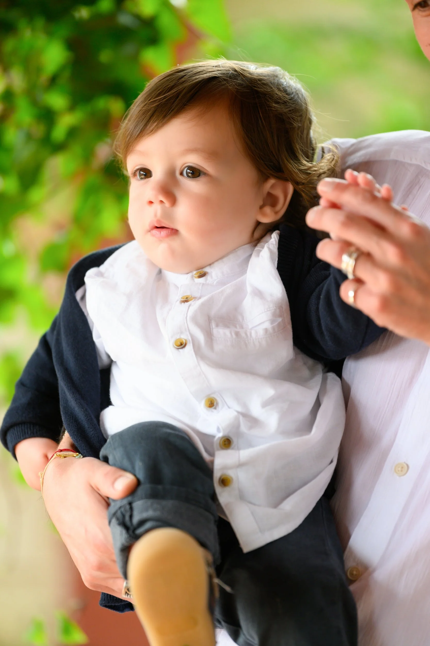 A young child with brown hair and big brown eyes, sitting on an adult's lap. The child is wearing a white shirt with gold buttons, a dark cardigan, and dark pants. The background shows a blurred outdoor setting with green foliage.
