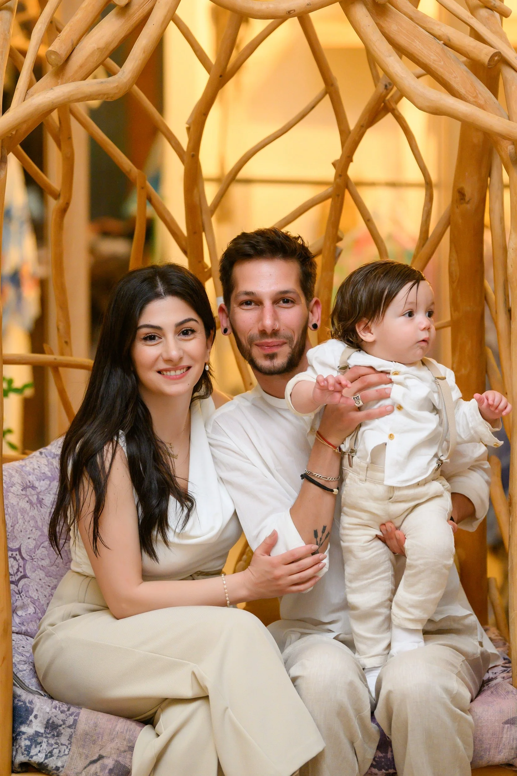 A family of three sitting on a purple cushioned seat in front of a wooden decorative backdrop. The woman has long dark hair and is smiling, wearing a white sleeveless top and beige pants. The man has short dark hair, a beard, earrings, and is holding