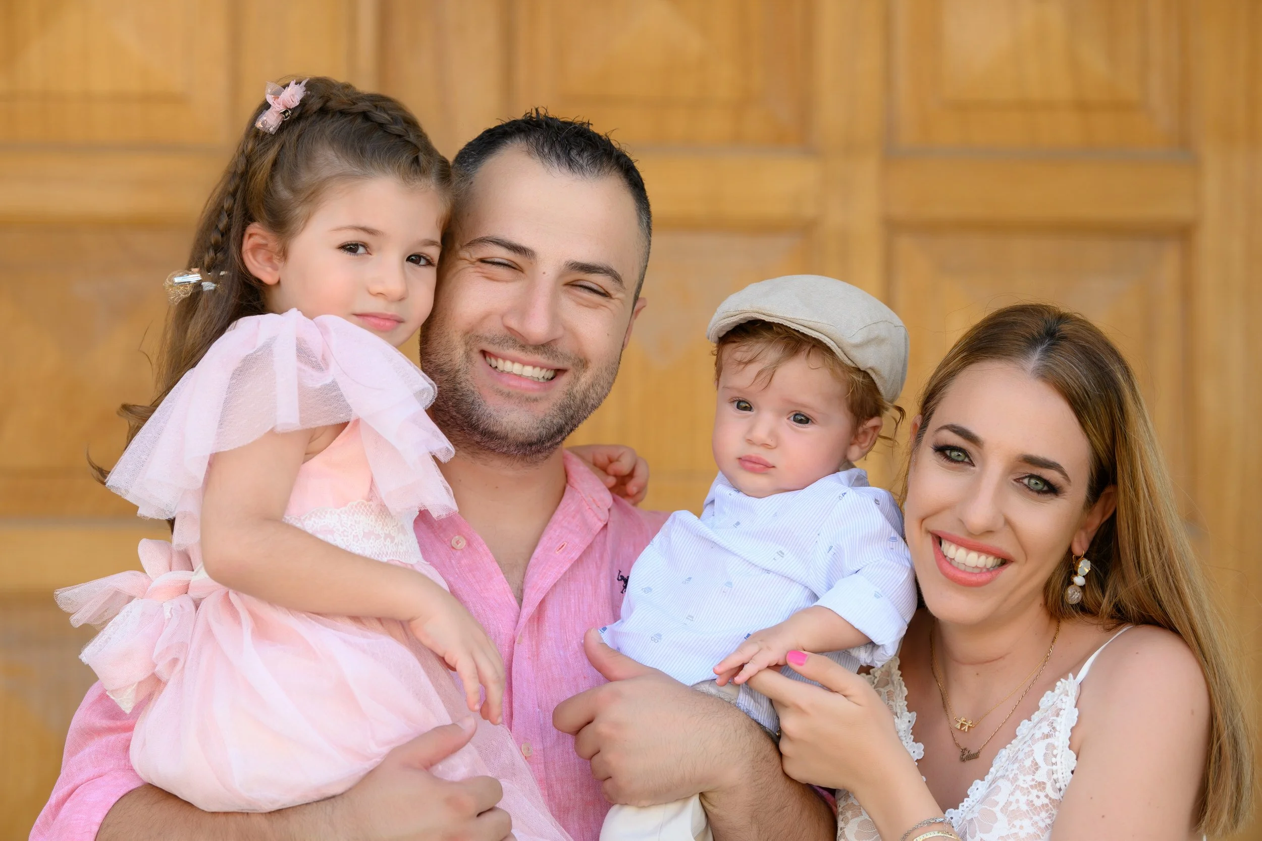 Happy family of four smiling and posing in front of a wooden background, with a father holding a young girl and a mother holding a baby boy.