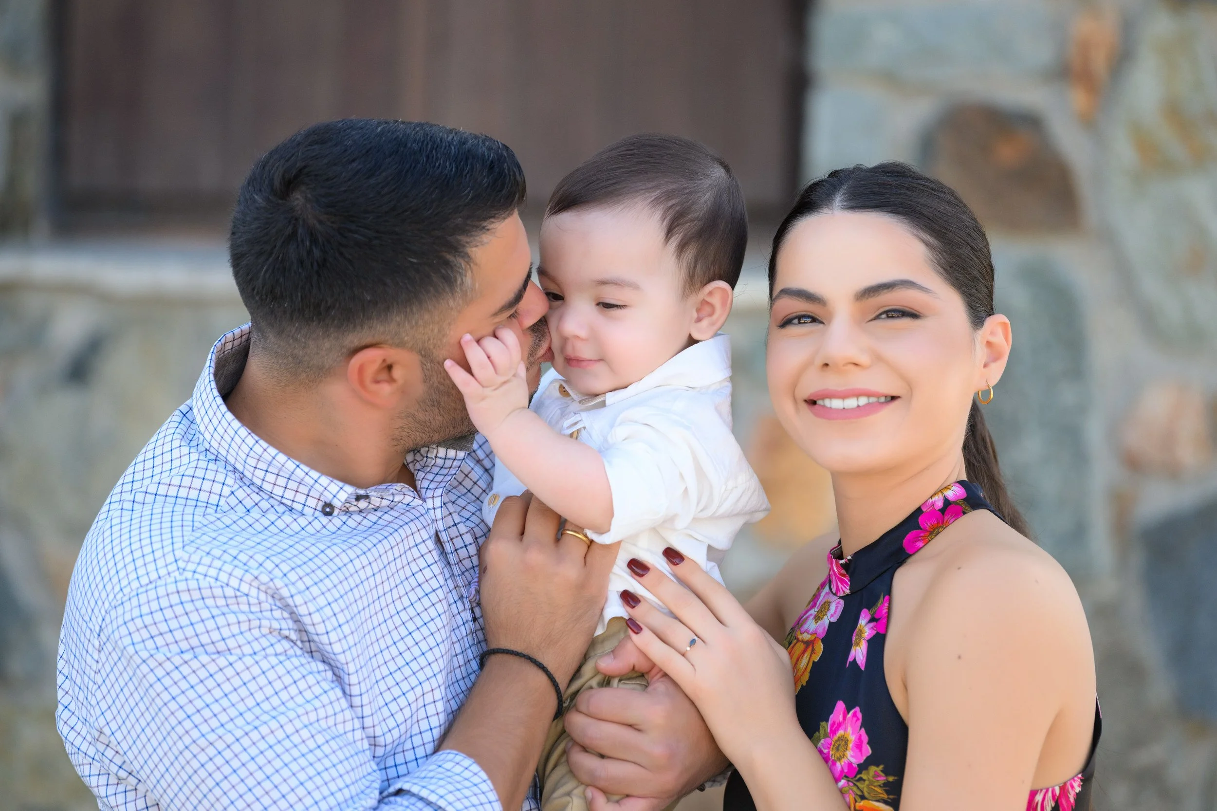 Three people, a man, a woman, and a young boy, outdoors in front of a stone wall. The man and boy are close, with the boy touching the man's face and the man smiling at him. The woman is smiling at the camera, holding the boy's waist.