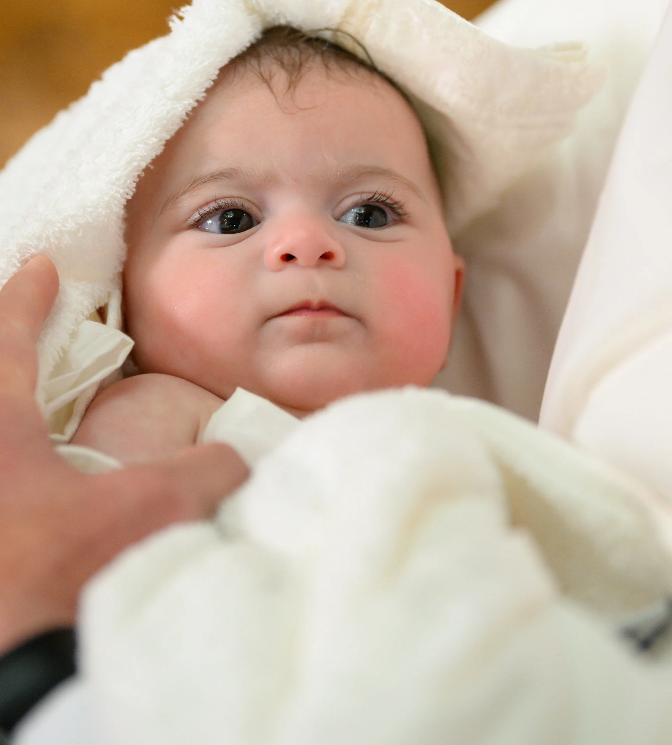 Close-up of a baby with blue eyes, wrapped in a white towel or blanket, with a hand gently touching its shoulder.
