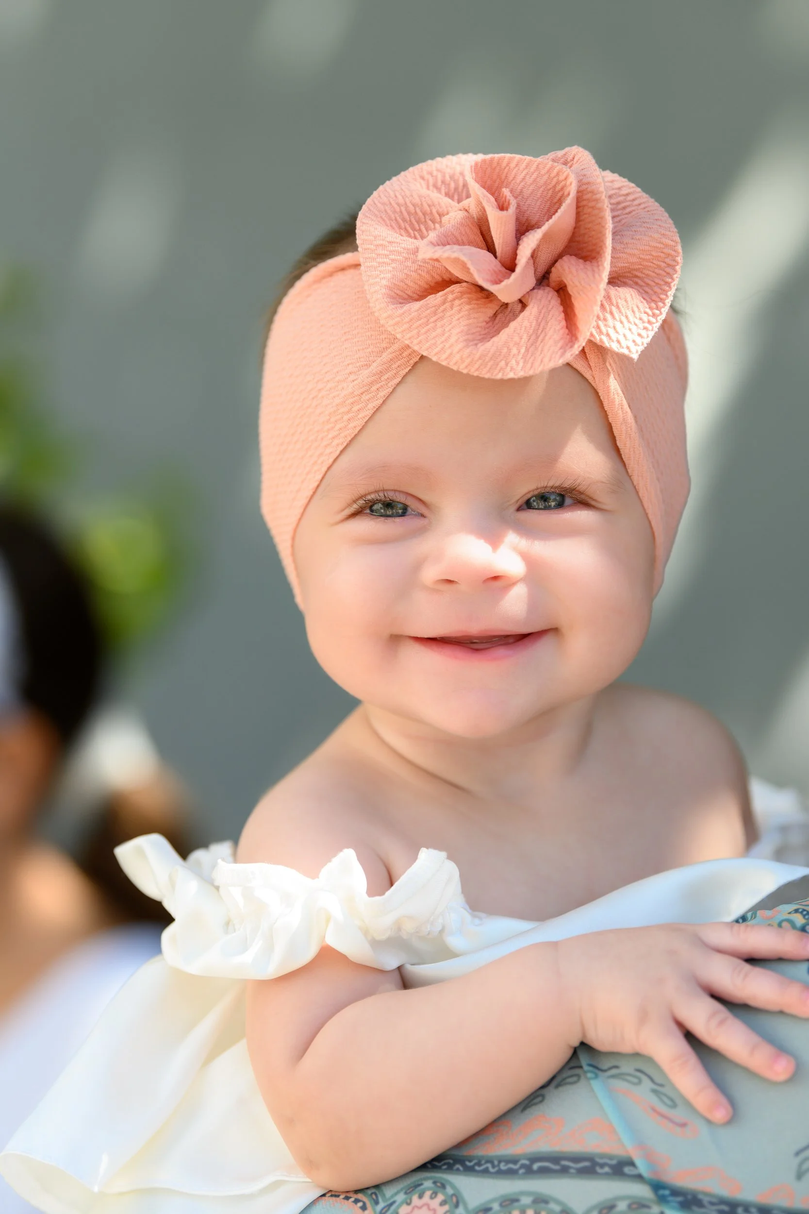 A smiling baby girl with blue eyes wearing a peach-colored headband with a large fabric flower on it, being held by someone outdoors.