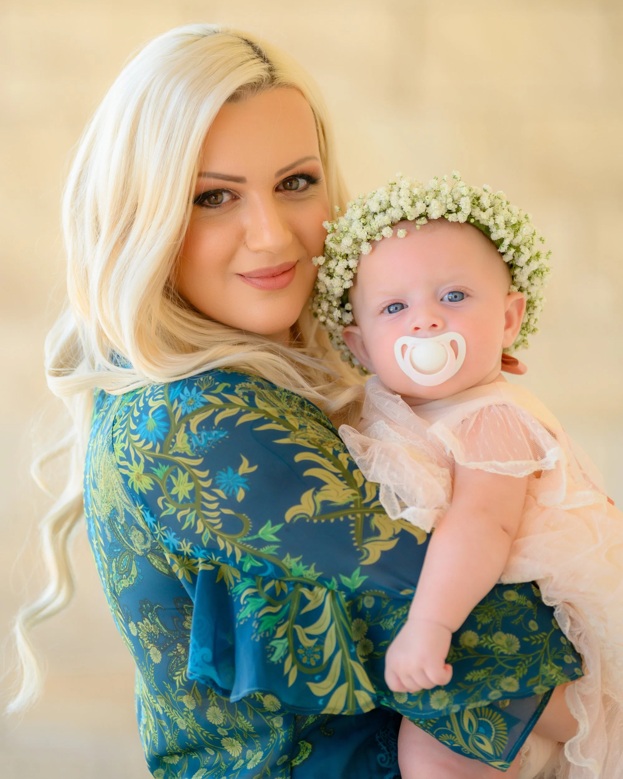 A woman with long blonde hair holding a baby girl with a pacifier and a floral crown, both looking at the camera. The woman is wearing a blue patterned dress.