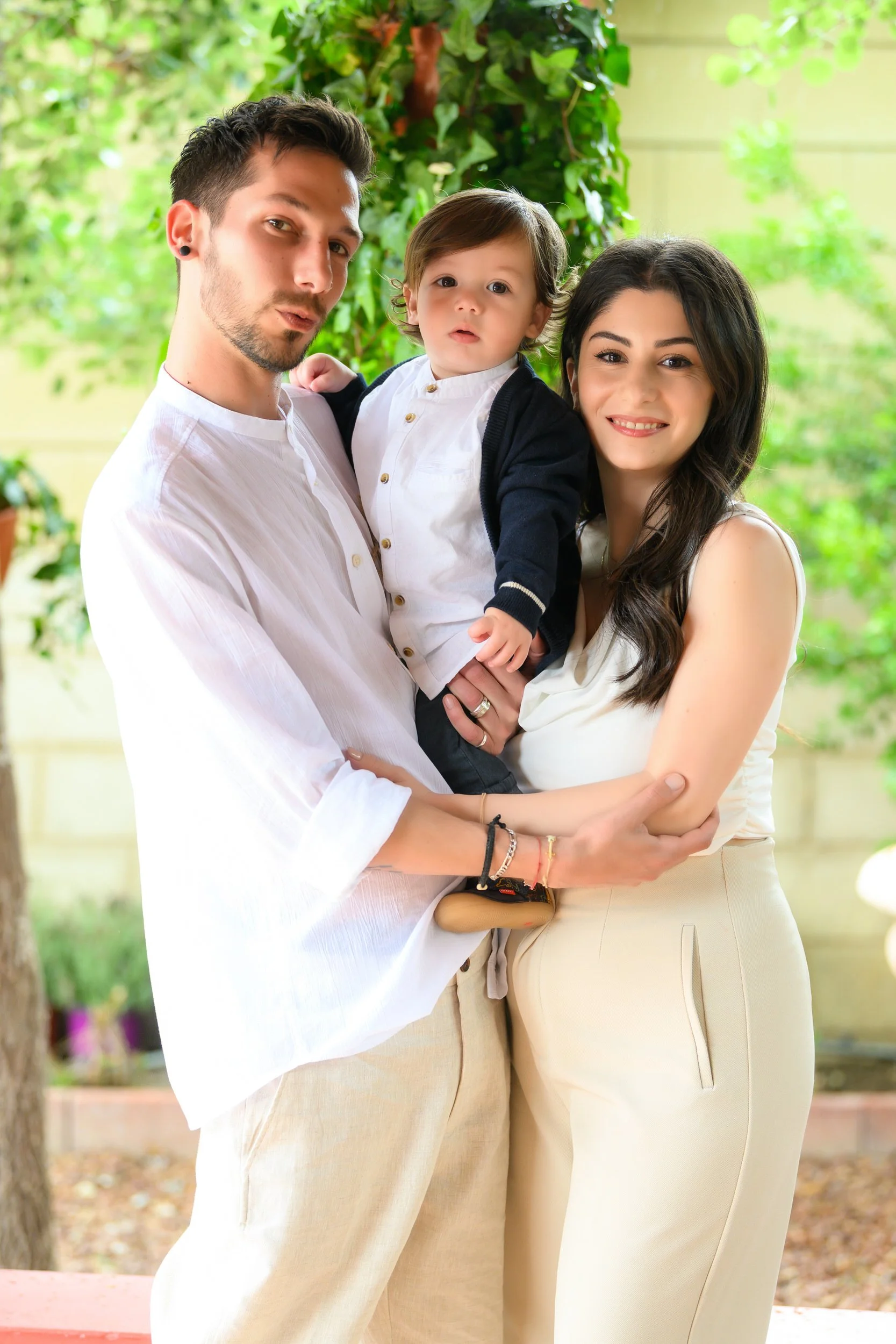 A family of three, with a man holding a young boy and a woman standing beside them, outdoors with green foliage in the background.