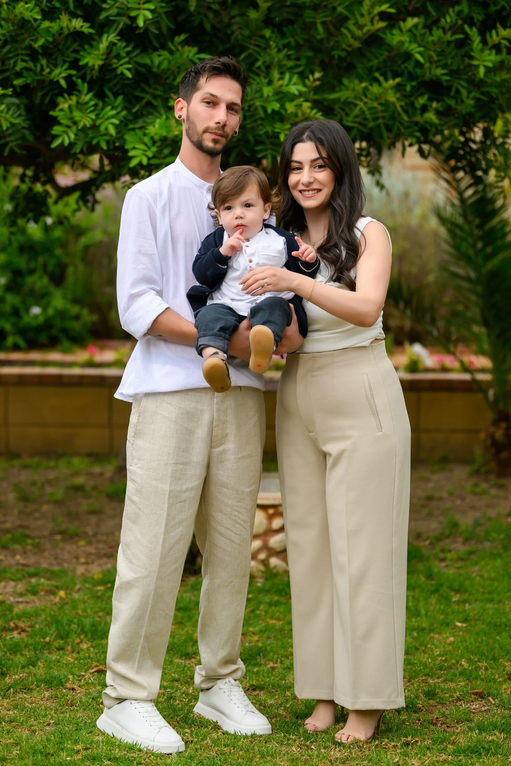 A young family of three standing outdoors in a garden. The man has dark hair, beard, and earrings, wearing a white shirt and beige pants. The woman has long dark hair, wearing a white top and beige wide-leg pants. They are holding a toddler boy dress