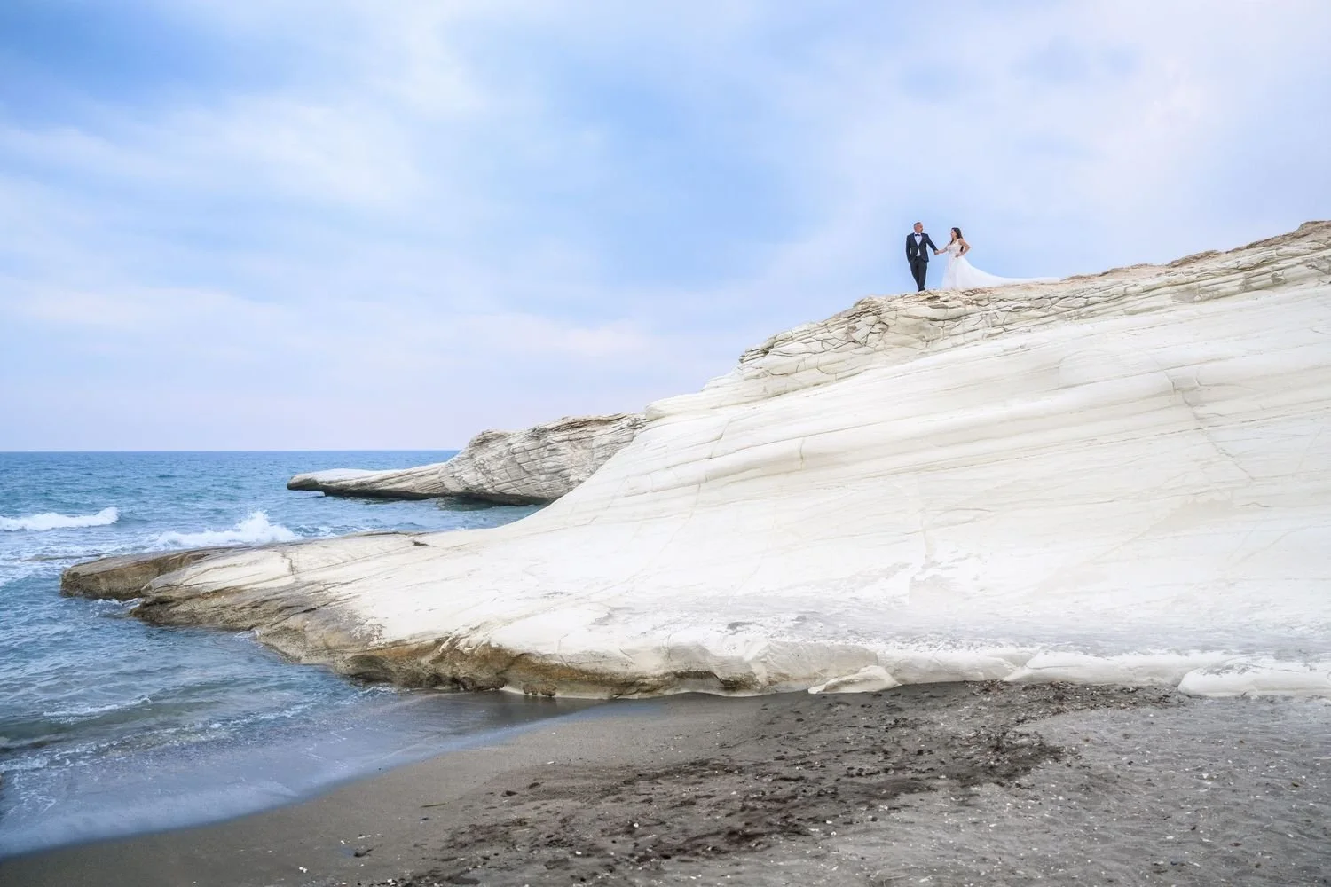 A bride and groom dressed in wedding attire holding hands on a white rock formation at the beach, with the ocean and partly cloudy sky in the background.