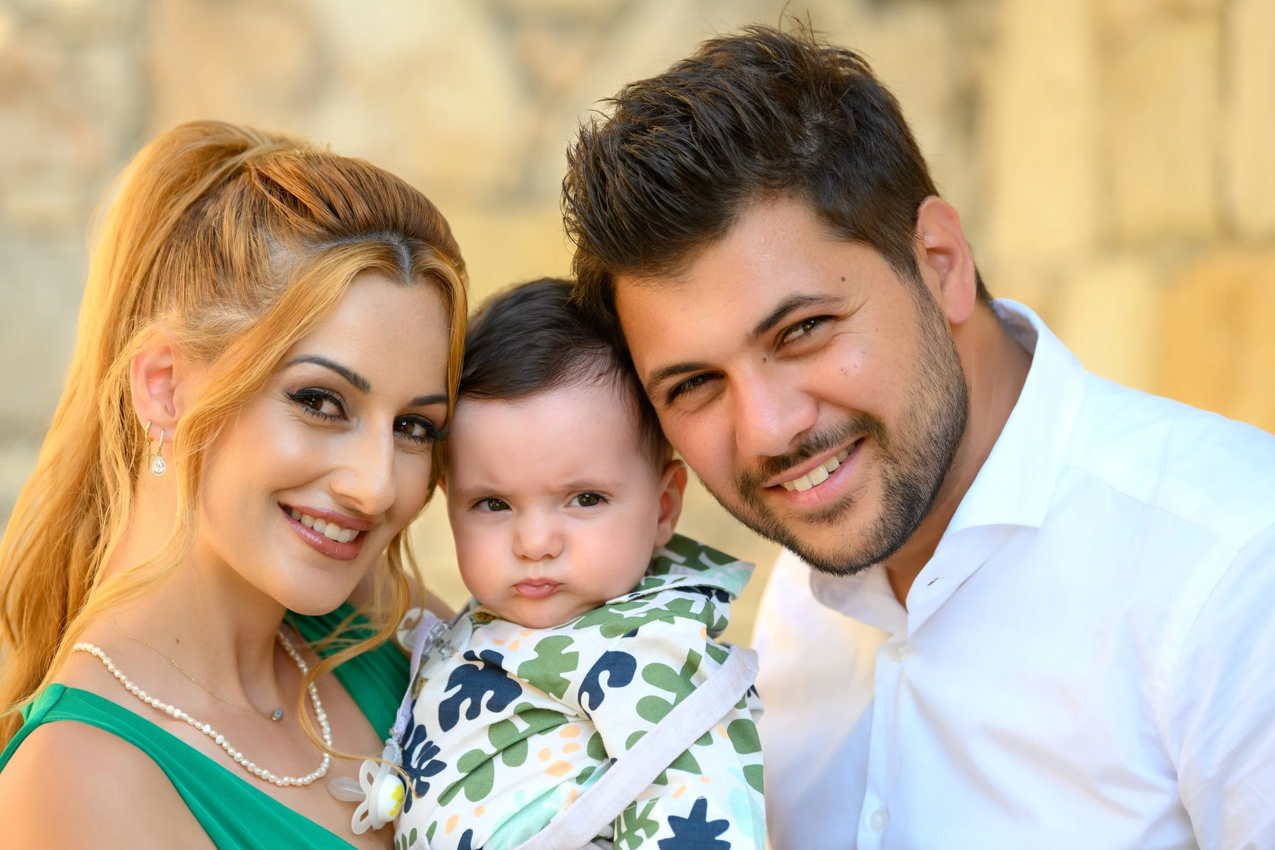 A smiling woman with blonde hair holding a young child with dark hair, and a smiling man near them. They are outdoors with a blurred background.