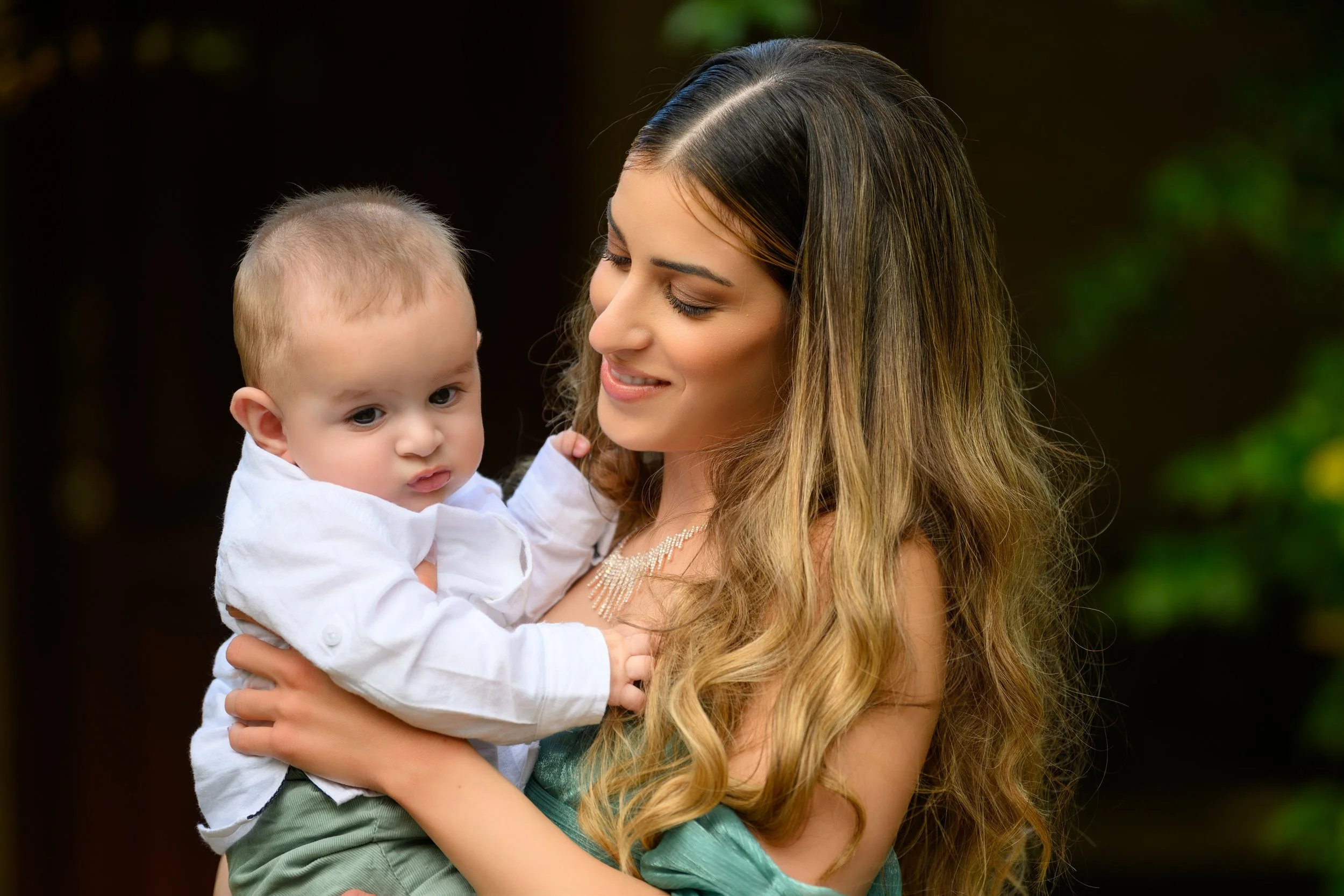 A woman with long, wavy blonde hair holding a young boy with short blonde hair outdoors, both looking at each other.