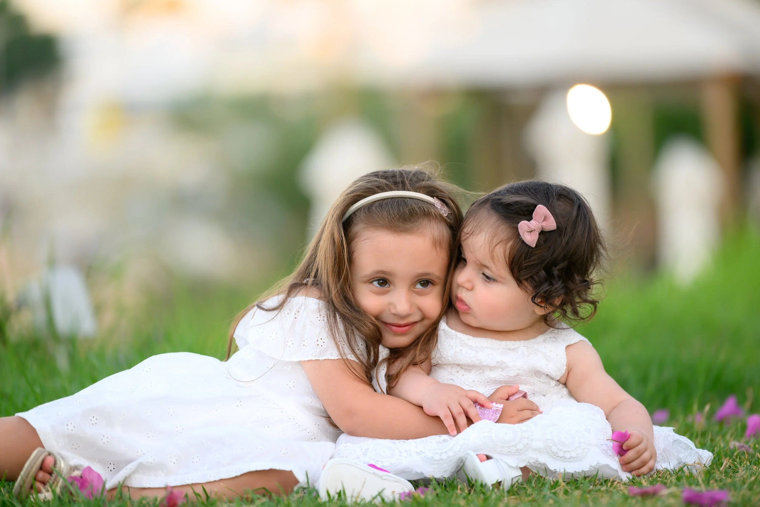 Two young girls in white dresses lying on the grass with pink flowers, in a blurry outdoor background.