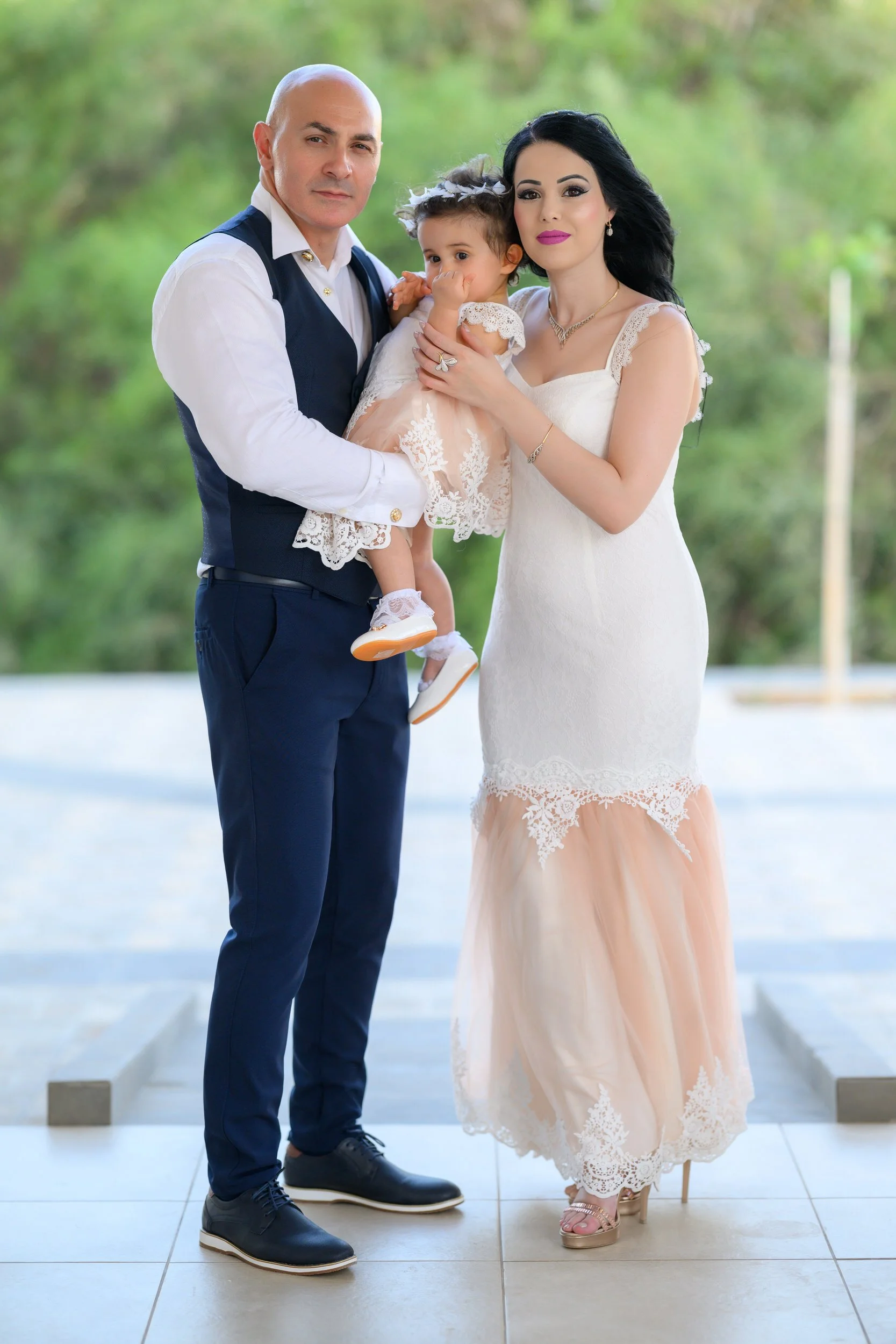 A family of three standing outdoors. The man is wearing a white shirt and navy vest and pants, holding a young girl dressed in a peach and white lace dress. The woman is wearing a white and peach lace dress with high heels, standing next to them with