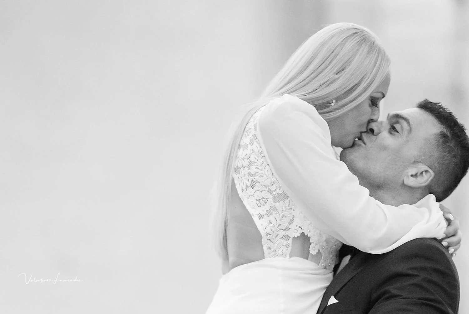 A black-and-white photo of a woman with long blonde hair, wearing a lace dress and leaning in for a kiss on the lips with a man in a tuxedo, who is holding her up and smiling.