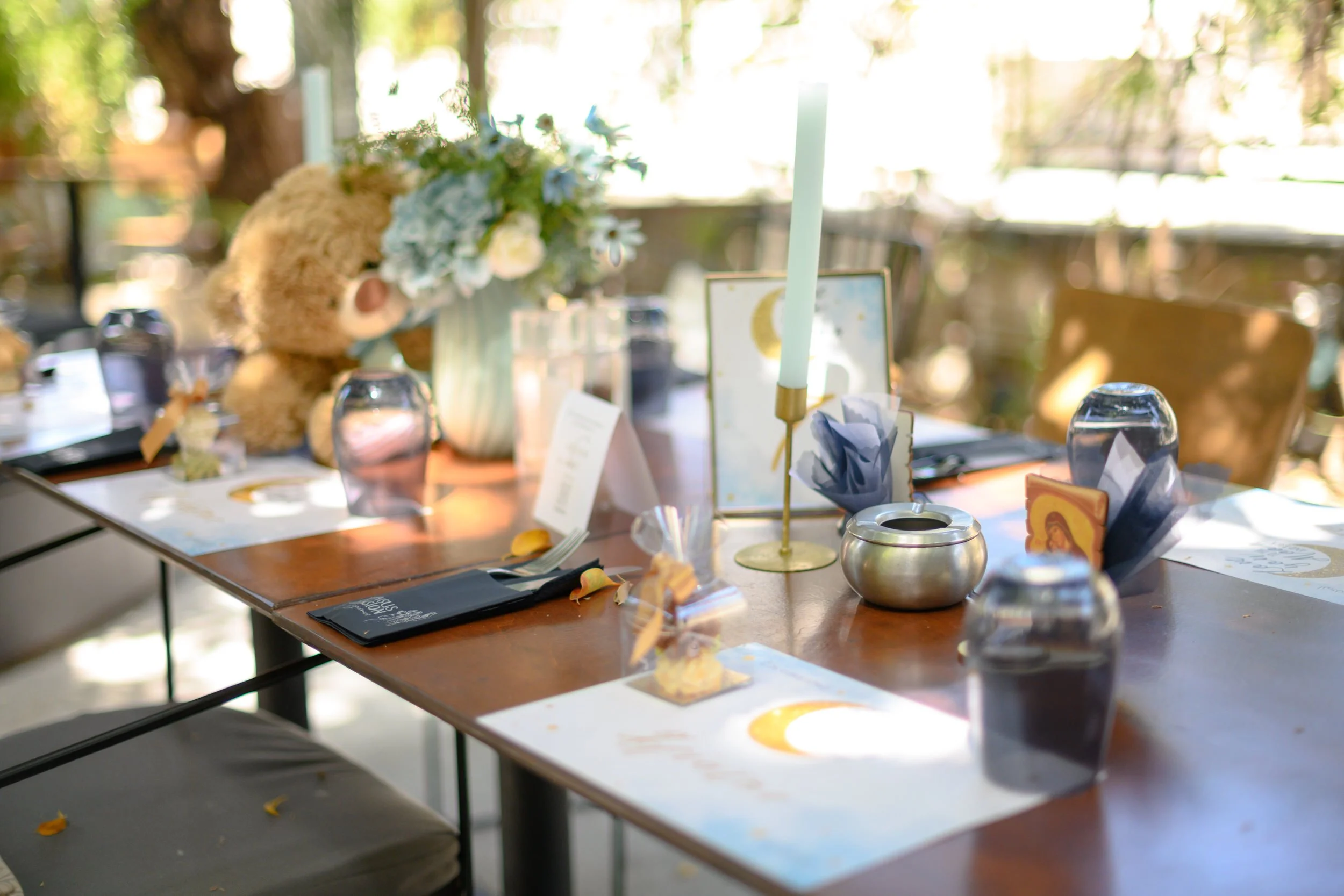 A dining table decorated with a floral centerpiece, candles, napkins, and celebration decorations, set outdoors with sunlight filtering through trees.