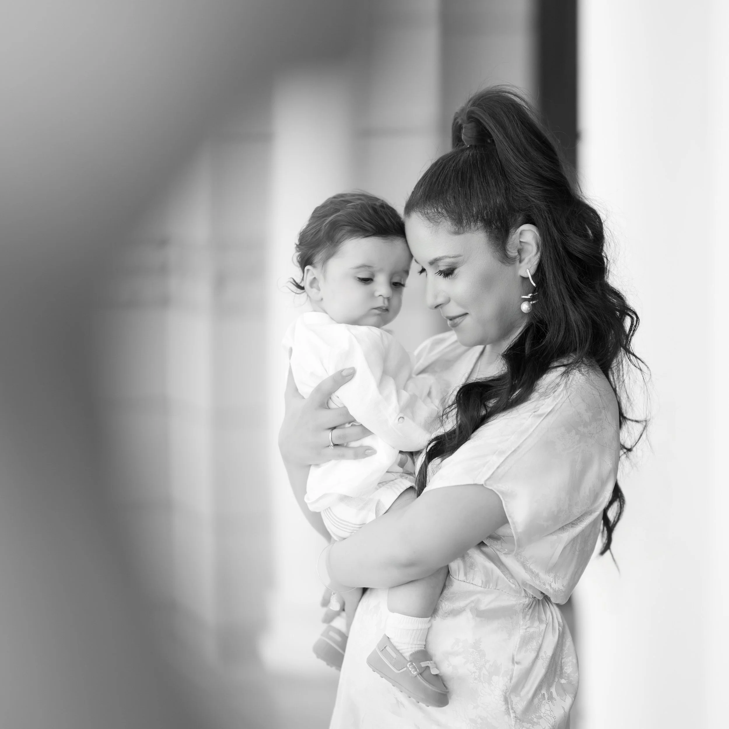 Black and white photo of a woman holding a young child close, looking down softly at the child, with a blurred background.