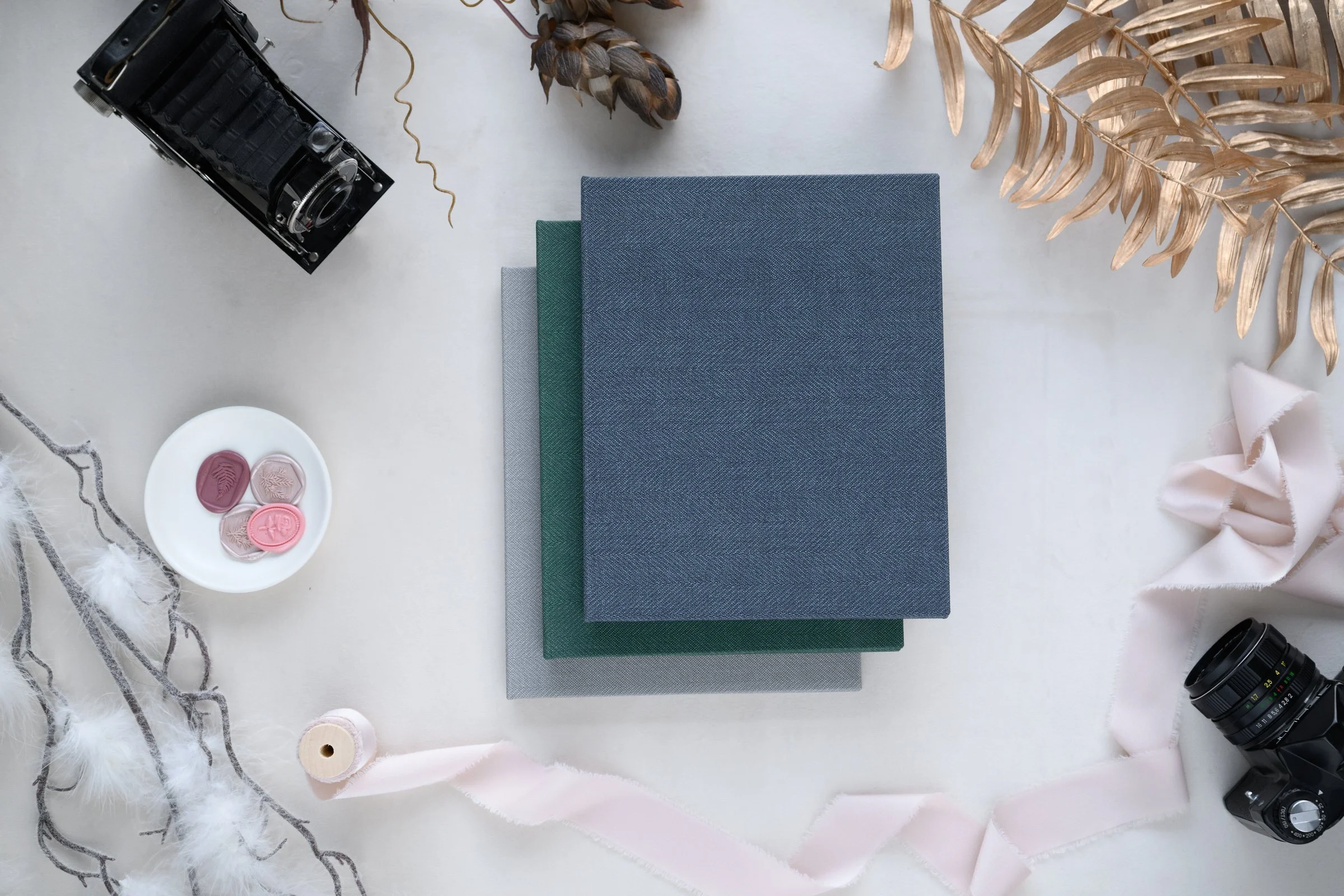 Flat lay of three fabric-covered usb folios in blue, green, and gray, surrounded by dried leaves, a roll of pink ribbon, a small white dish with pink decorative stones, and two vintage cameras on a light-colored surface.