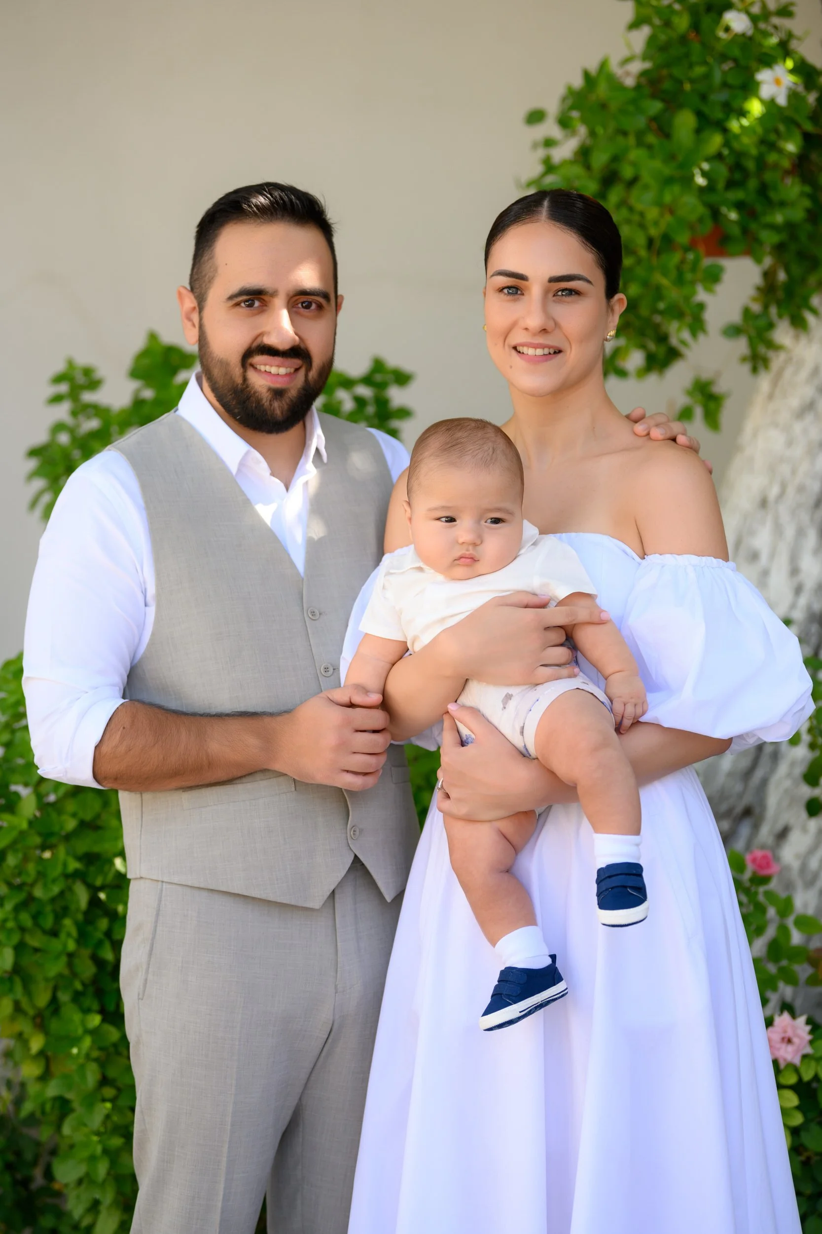A family of three standing outdoors with greenery. The man has a beard and is wearing a white shirt with a light beige vest. The woman has dark hair in a bun and is wearing a white off-the-shoulder dress. They are holding a baby boy in a white shirt 