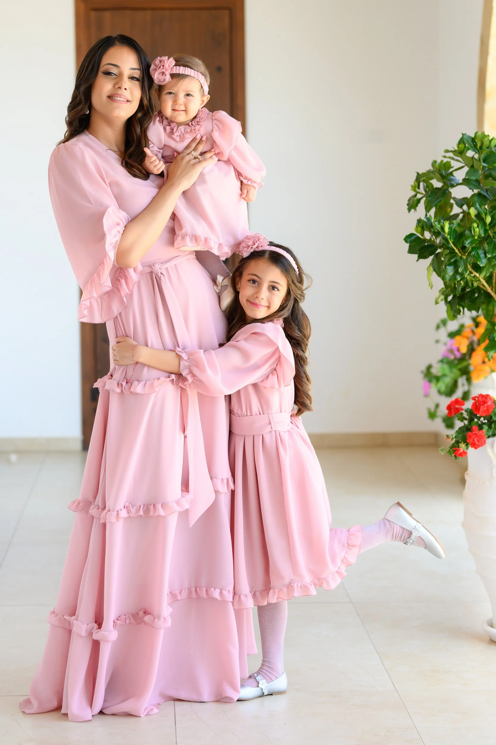 A woman with two young girls, all dressed in matching pink outfits, standing indoors near a door and a potted plant. The woman is holding a baby while the older girl hugs her legs, smiling at the camera.