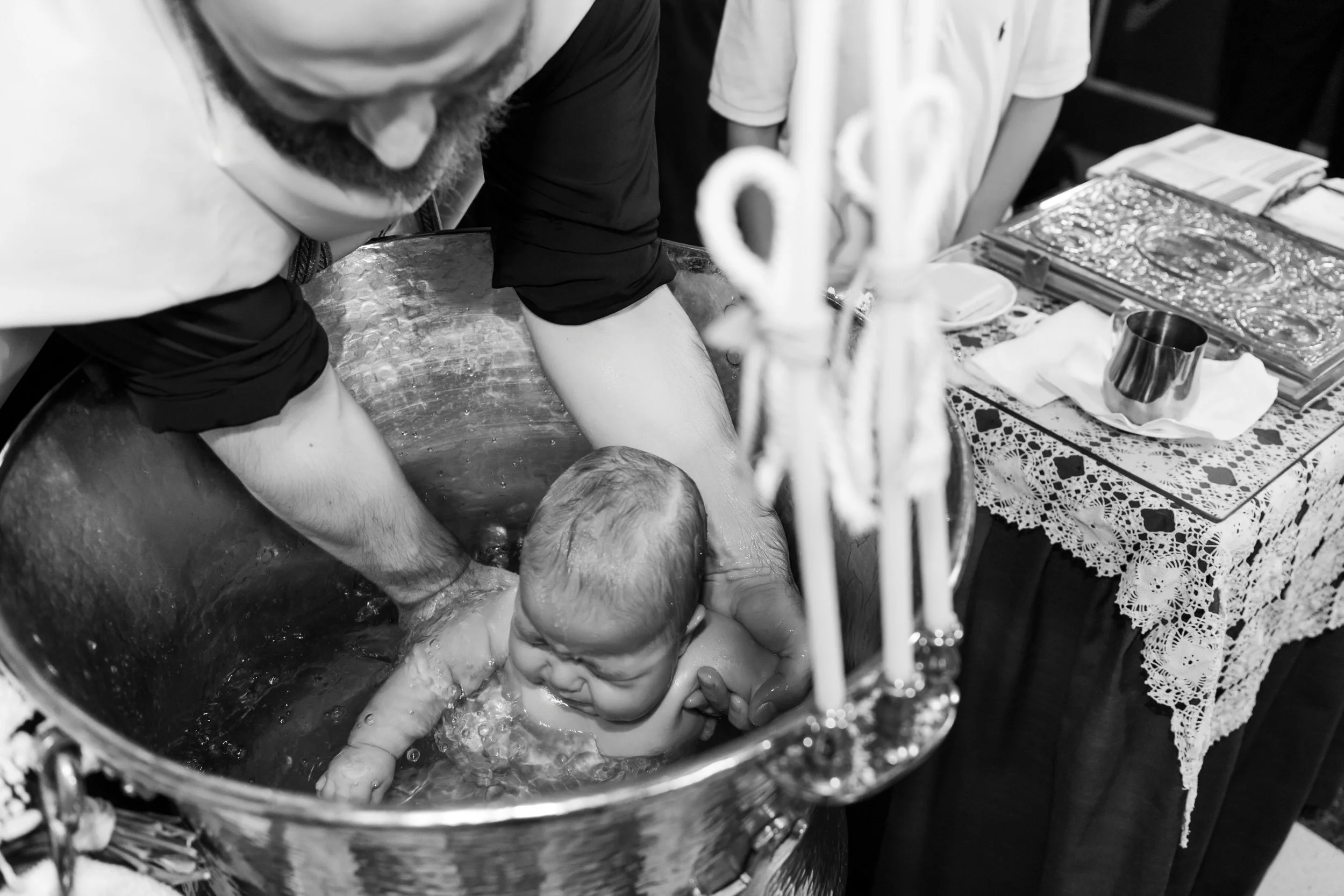 Child being baptized in a large metal baptismal font, assisted by an adult, with baptismal supplies and religious items on a table nearby.