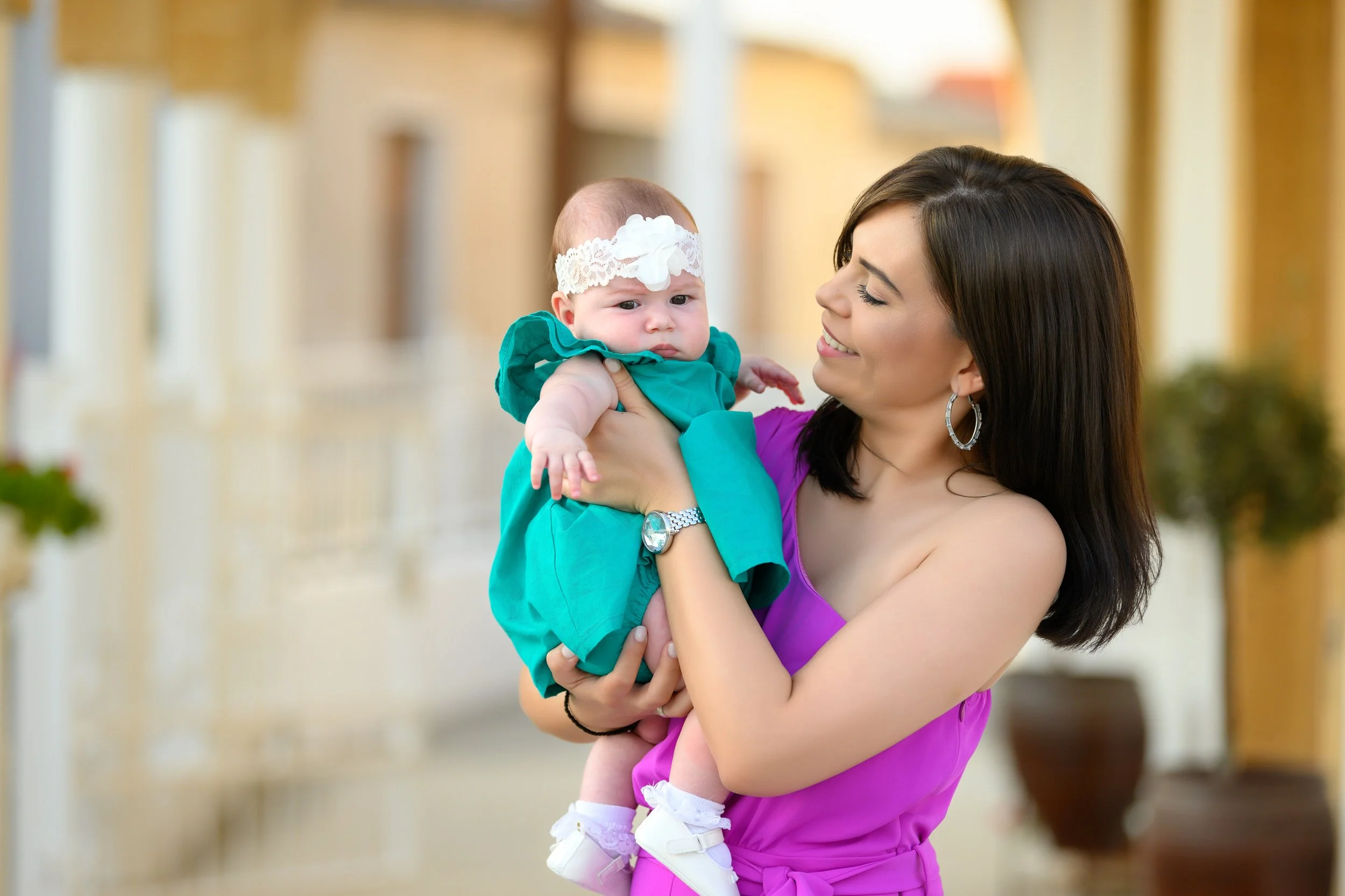 A smiling woman holding a baby girl dressed in a teal dress with a white lace headband, outdoors with a blurred background.