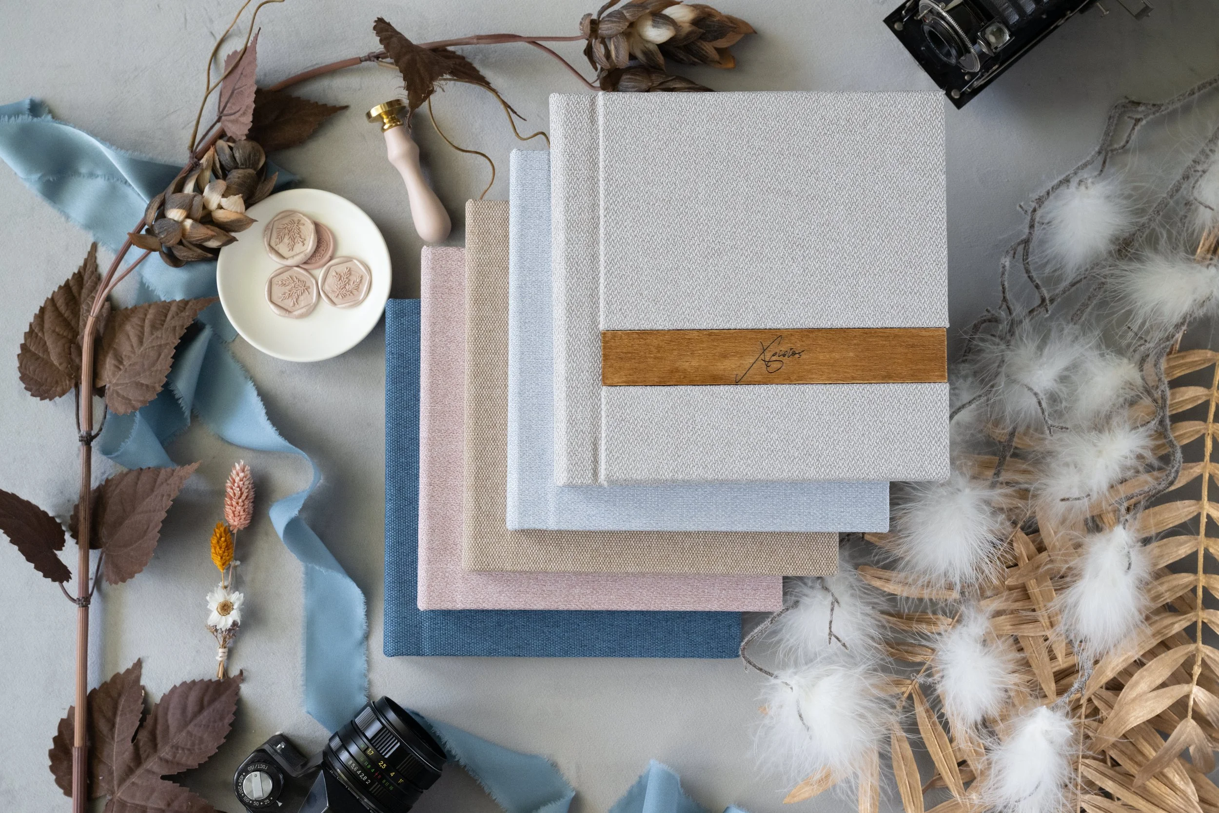 Arrangement of lay flat albums in neutral pastel tones, a small dish with wax seals, dried flowers, a vintage camera, and decorative grass and feathers on a light gray surface.