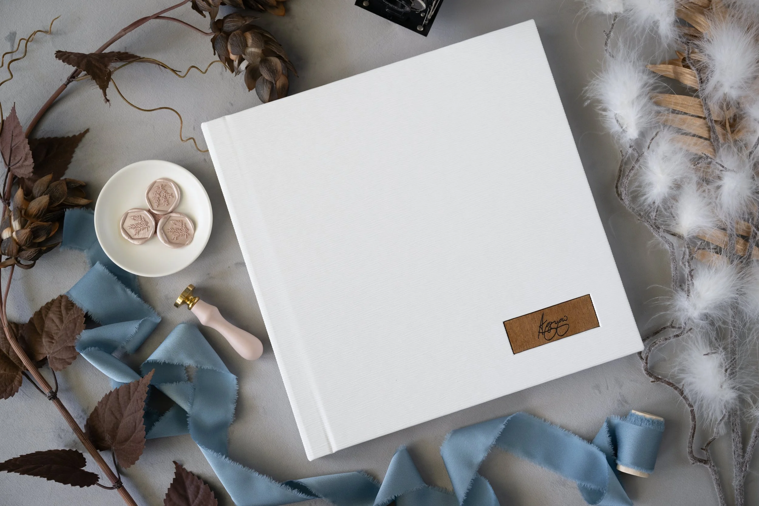 A white photo album with a leather tag on the cover, surrounded by dried leaves, soft blue ribbon, white feathers, and small hexagonal cookies on a plate.