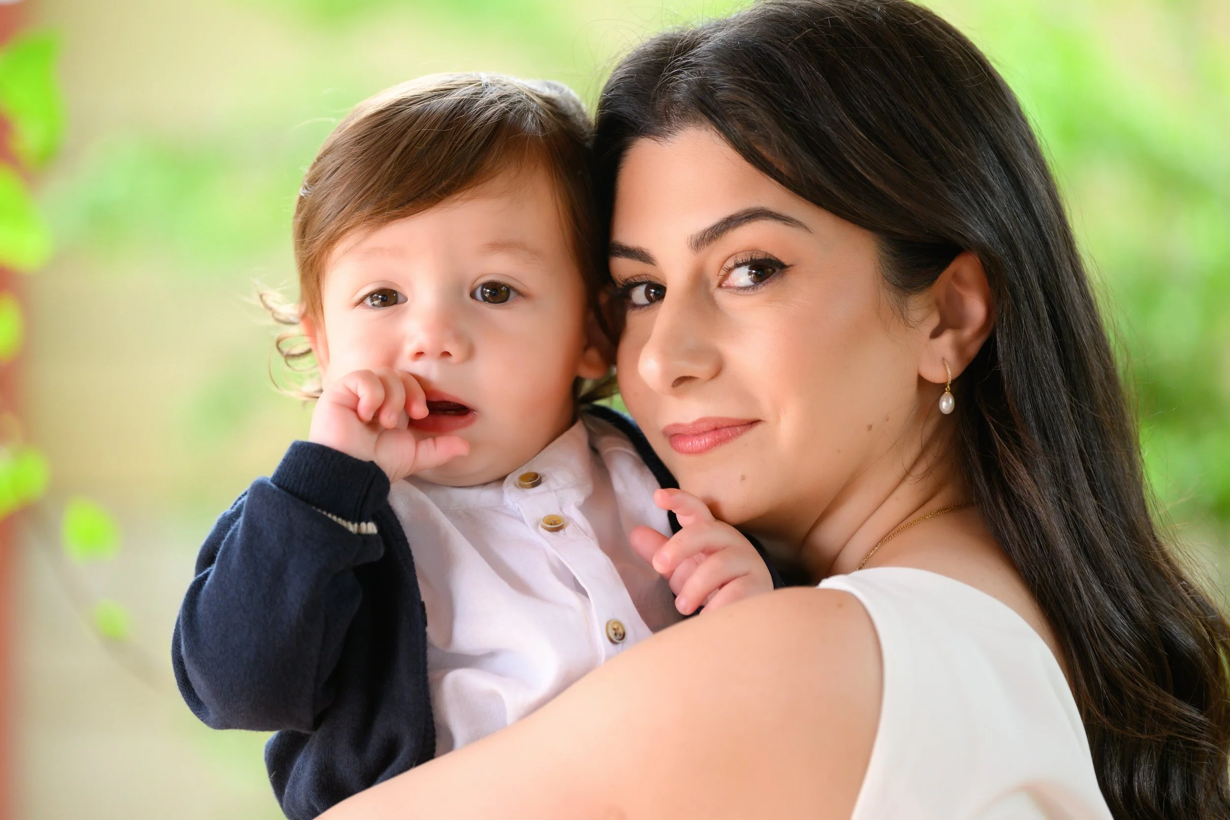 A young woman with dark hair and pearl earrings holding a toddler with light brown hair and big eyes, both looking at the camera in a natural outdoor setting.
