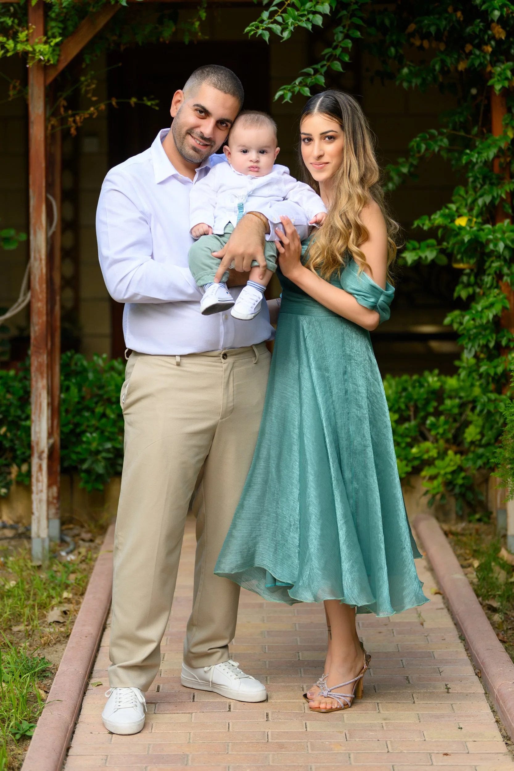 A family of three standing on a brick pathway outdoors, with greenery in the background. The man is wearing a white shirt and beige pants, holding a baby. The woman is in a green dress with long wavy hair, standing next to them.