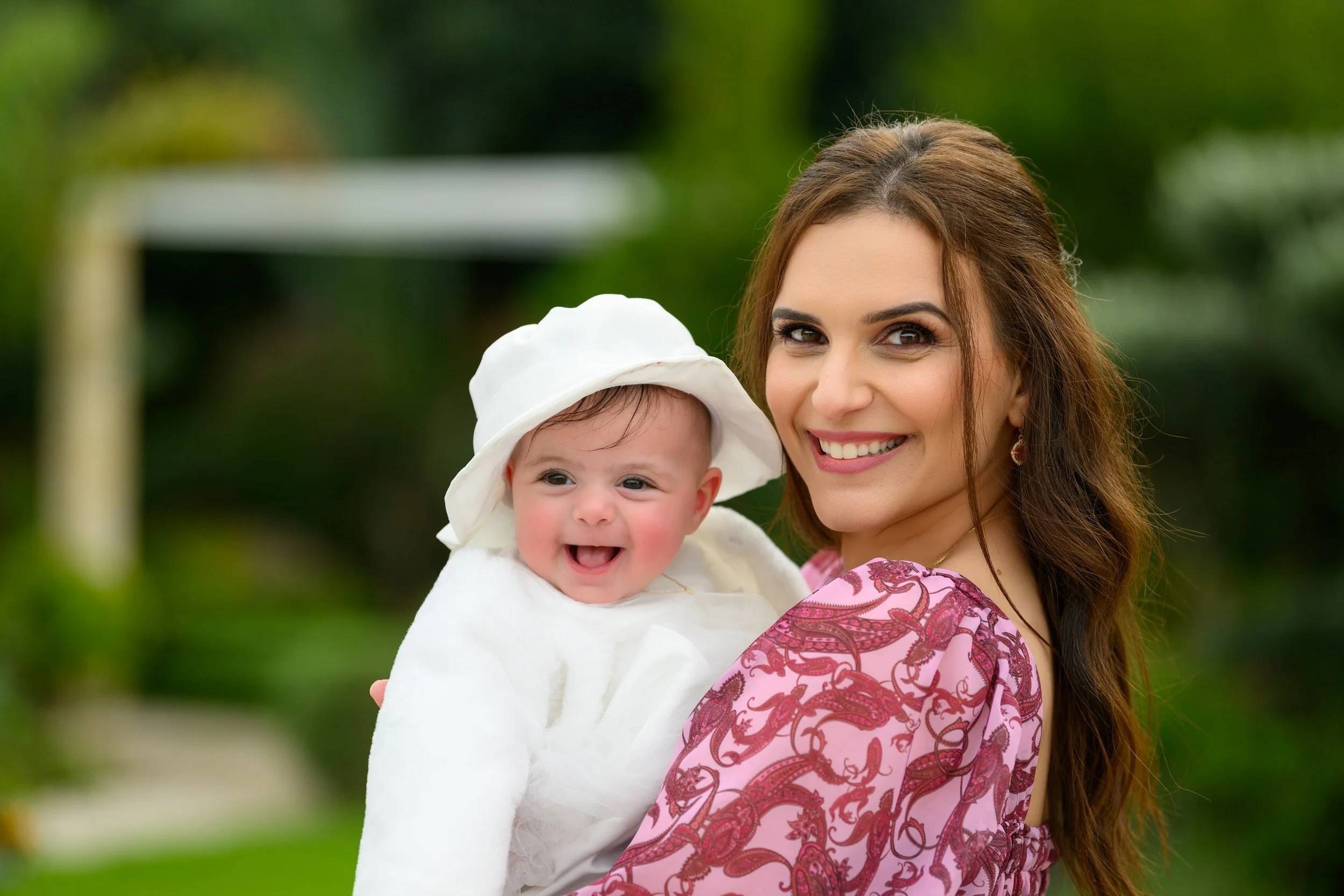 A smiling woman holding a happy baby girl dressed in white with a white hat outdoors.