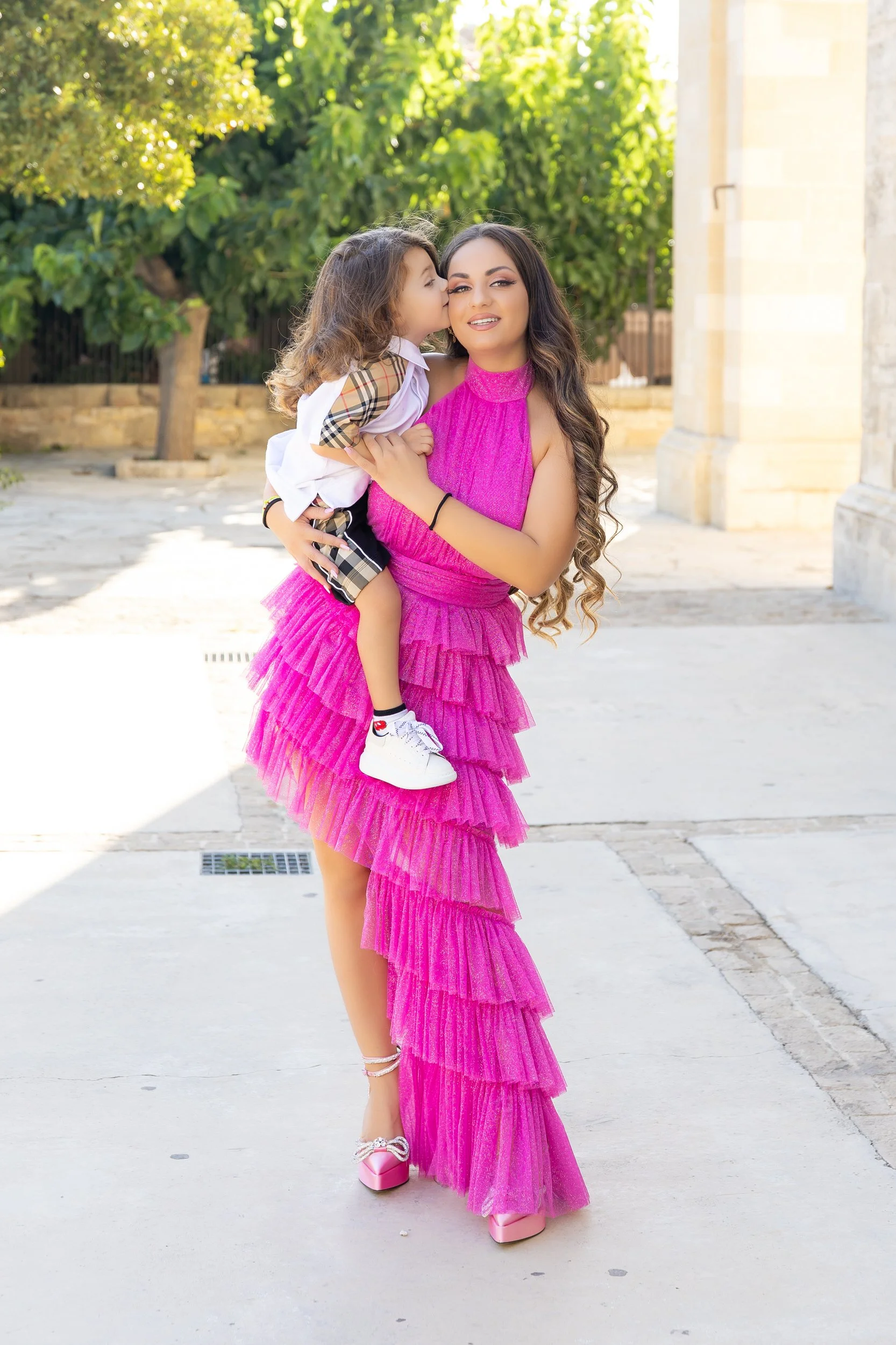 Young woman in a bright pink ruffled dress lifting a small child, who is giving her a kiss on the cheek, outdoors with trees and stone building in the background.