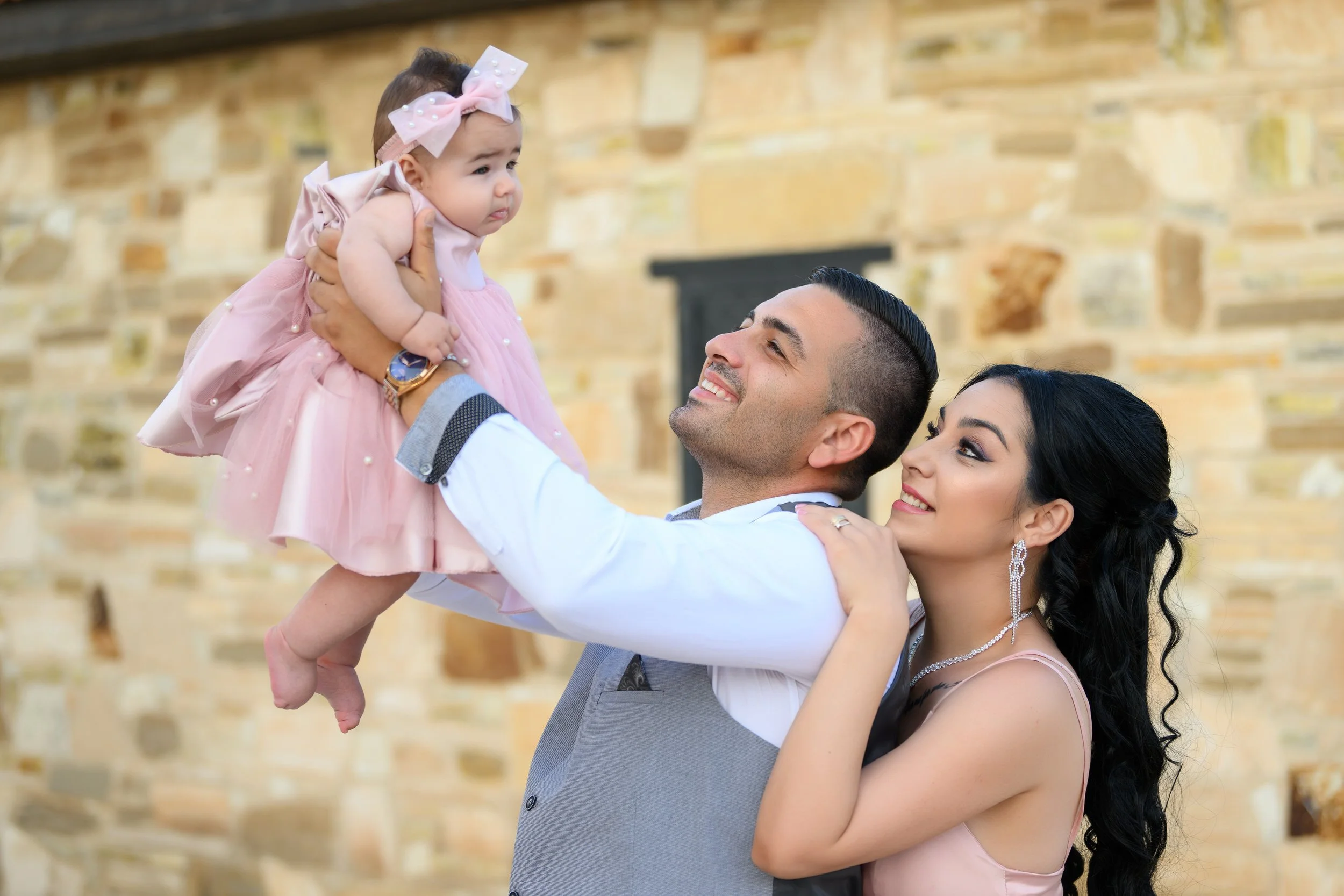 A man and woman smiling and looking at a baby girl they are holding up. The man is wearing a suit vest and the woman has long black hair and is dressed in a pink gown. The baby girl is dressed in a pink dress with a pink bow headband.
