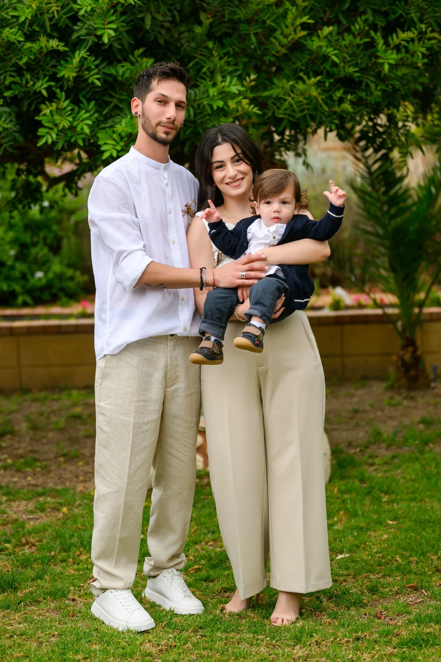 A happy family of three standing outdoors in front of green foliage. The woman is holding a young child, and the man stands beside them with one arm around the woman. The child is making a gesture with his hands. Both adults are smiling.