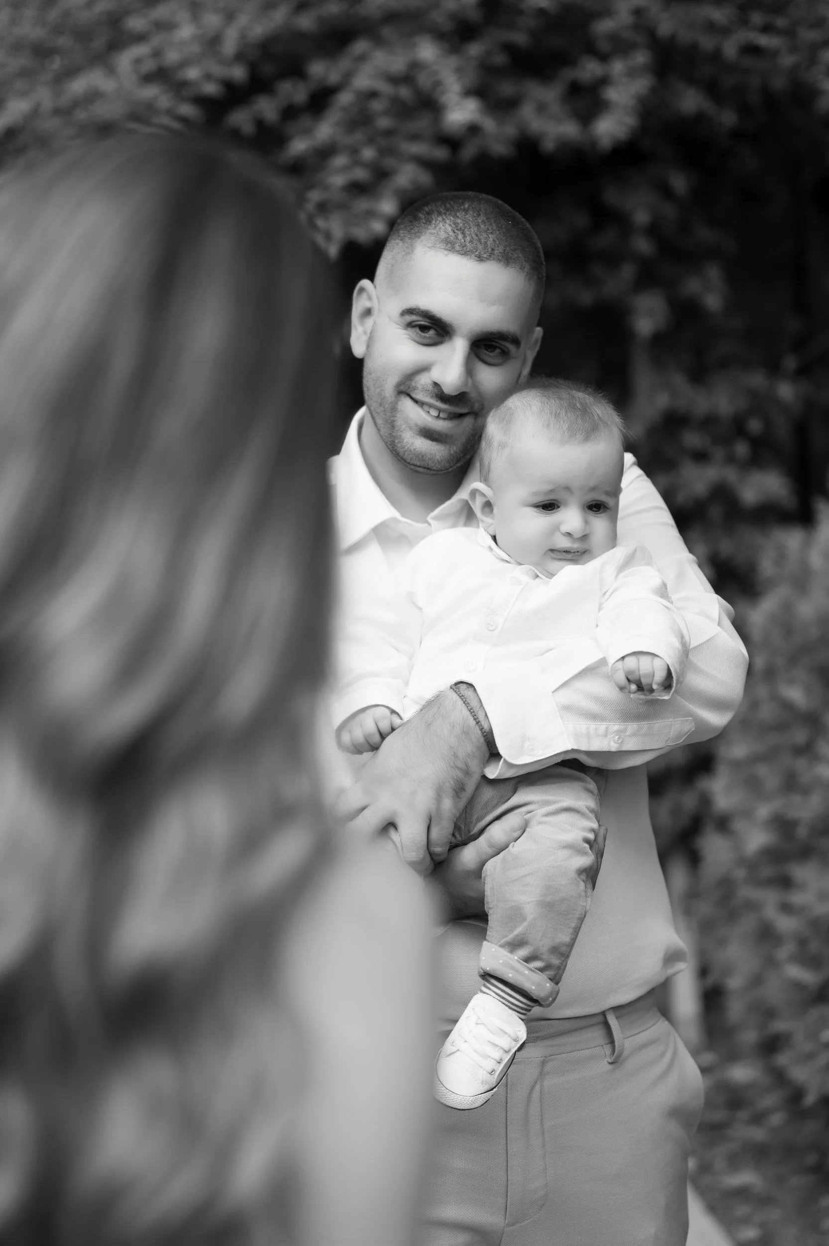 A man holding a young child and a woman in the foreground, with the woman facing away, outdoors with trees in the background.