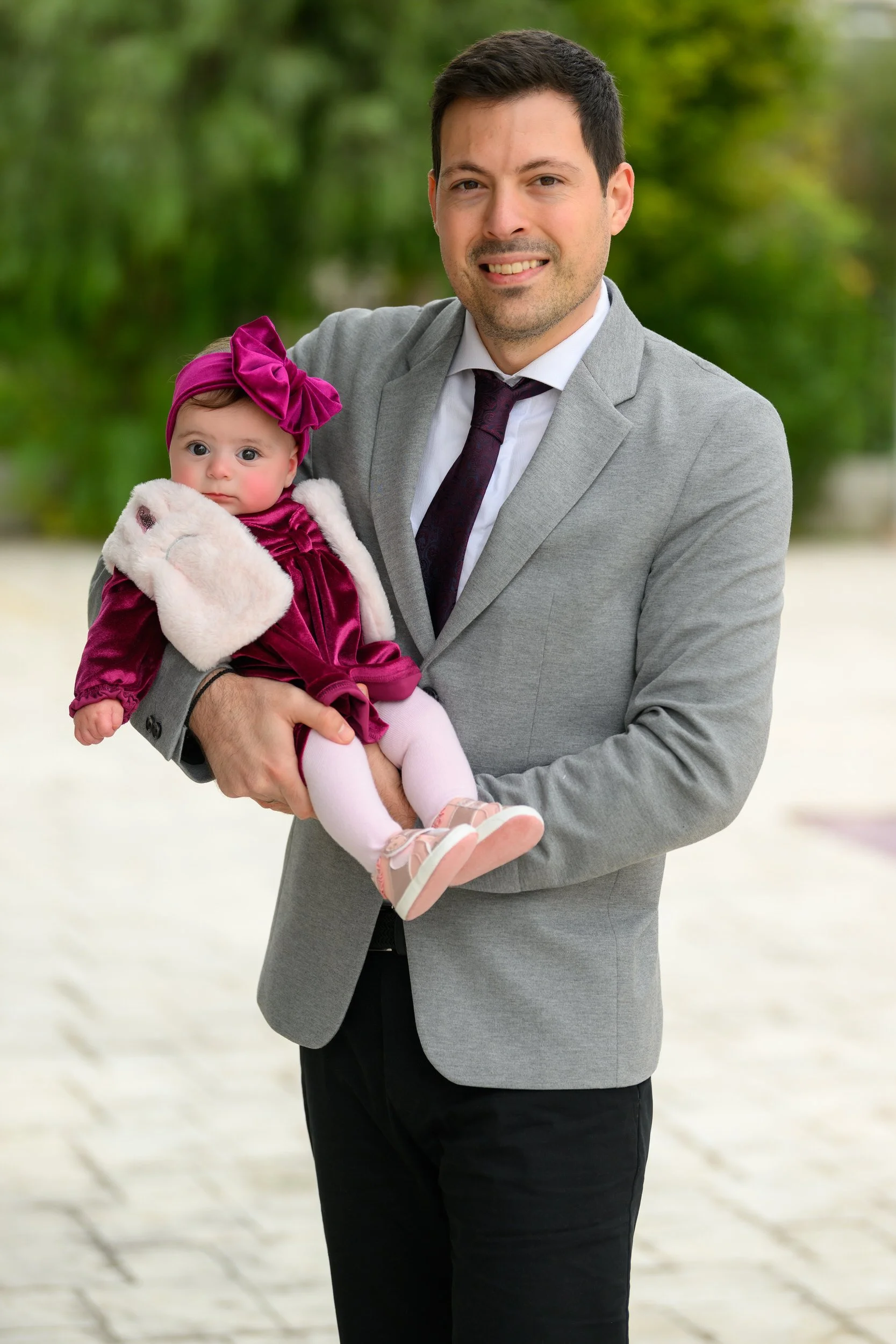 A man in a gray suit holding a baby girl dressed in a magenta velvet dress and headband, with white fur gloves and pink shoes, outdoors with green trees in the background.