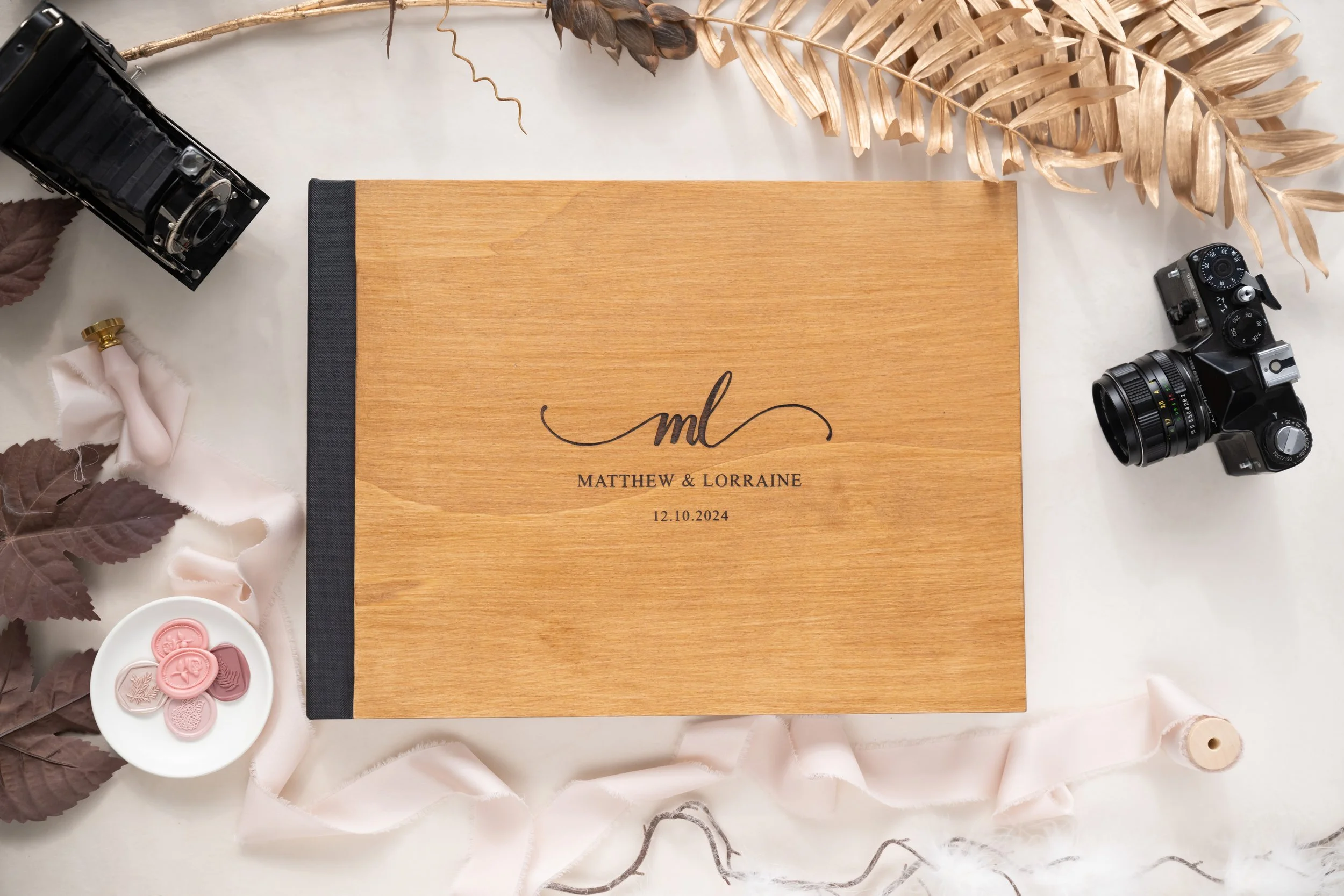 A wooden photo album with on the cover to the left, the initials 'ml' and the names 'Matthew & Lorraine' with the date '12.10.2024'. Surrounding the album are various vintage cameras, a white dish with pink wax seals, dried leaves, and a piece of pin
