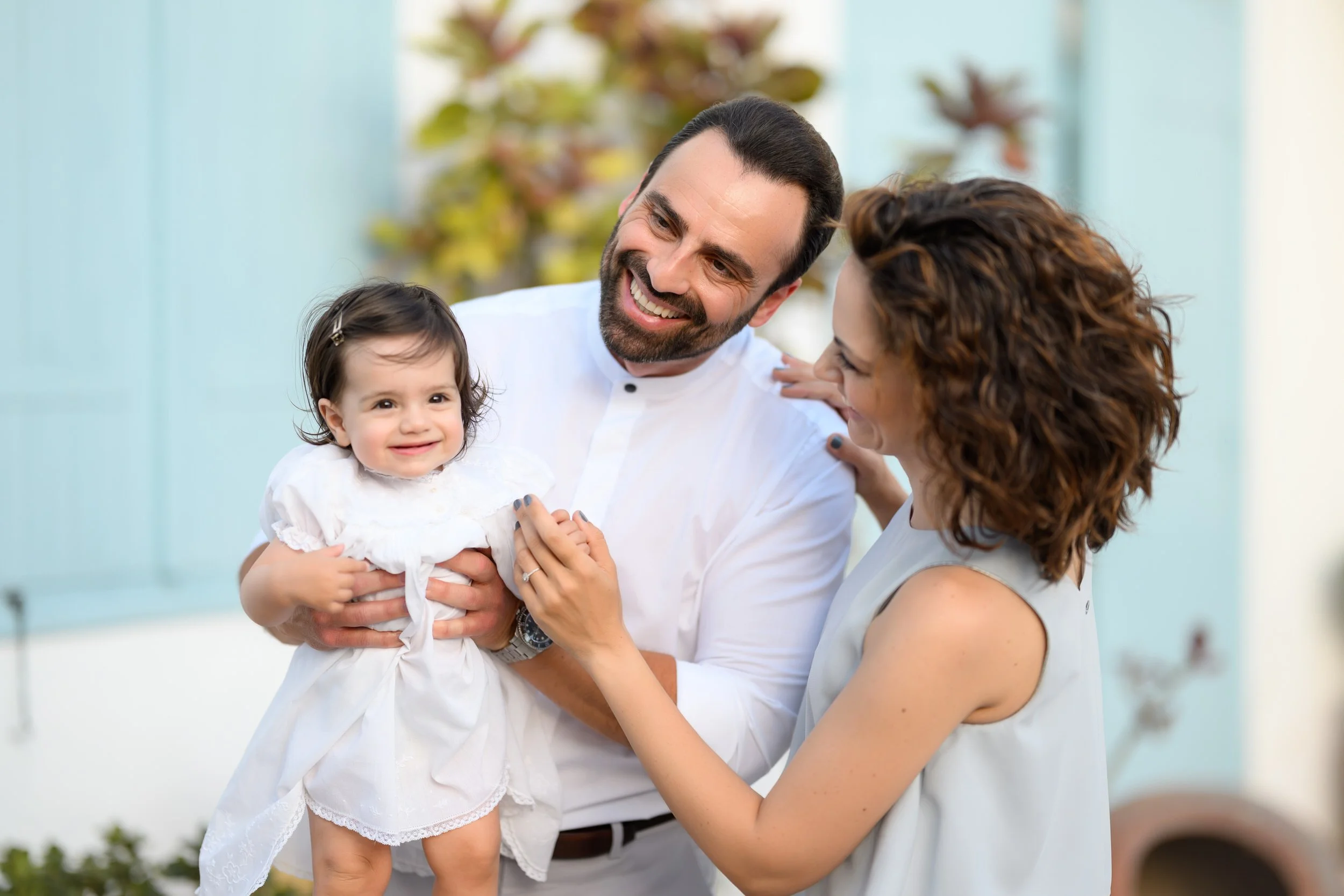 A happy family with a young girl in white dress, smiling while being held by a man and a woman, outdoors with blurred trees in the background.