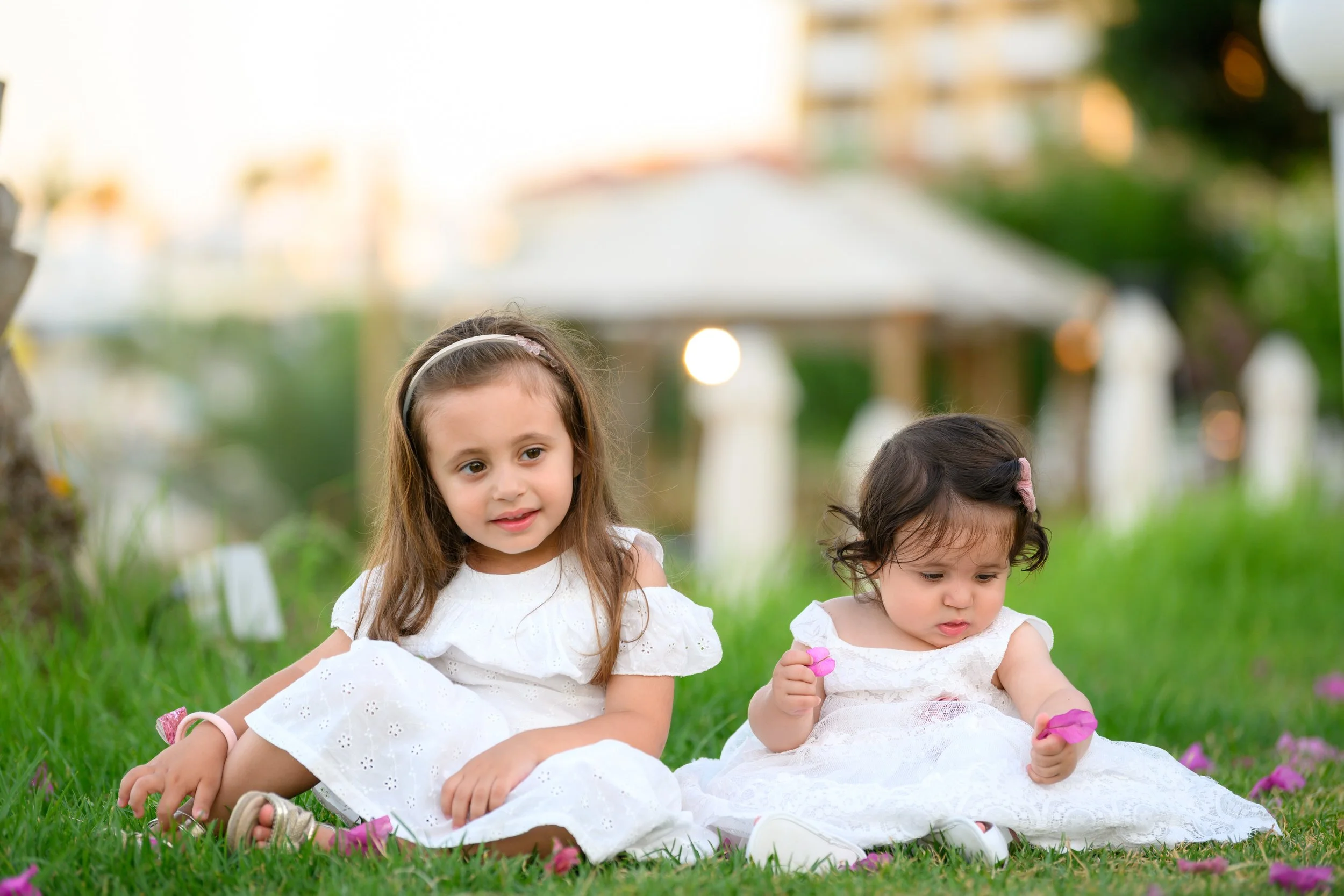 Two young girls sitting on grass in a park, wearing white dresses, with trees and a gazebo in the background.