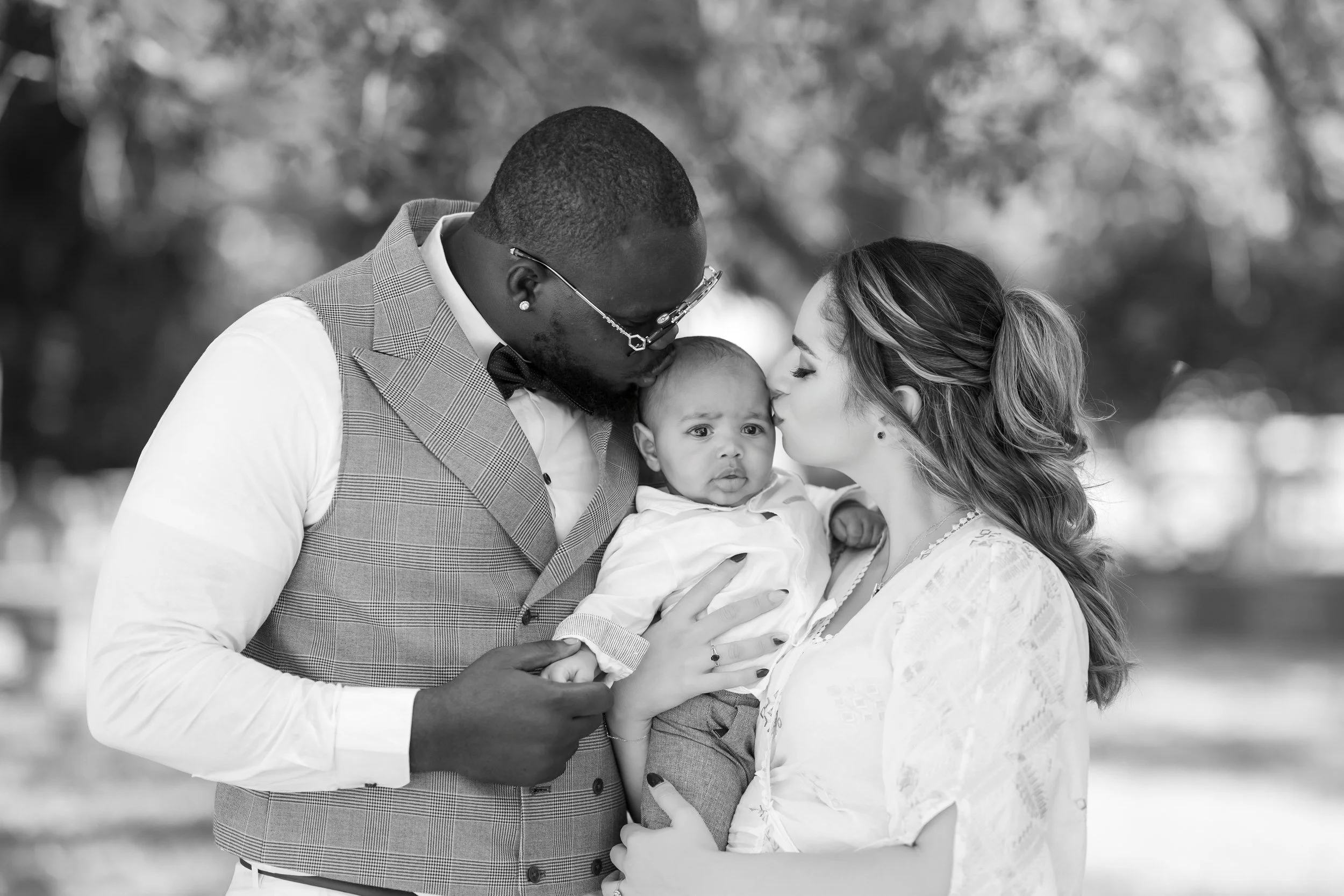 A black man and a white woman kiss a baby on the head outdoors. The man is wearing glasses, a checkered vest, and a bow tie. The woman has long, wavy hair and is wearing a light-colored dress. The baby looks confused and is held by the woman, with th
