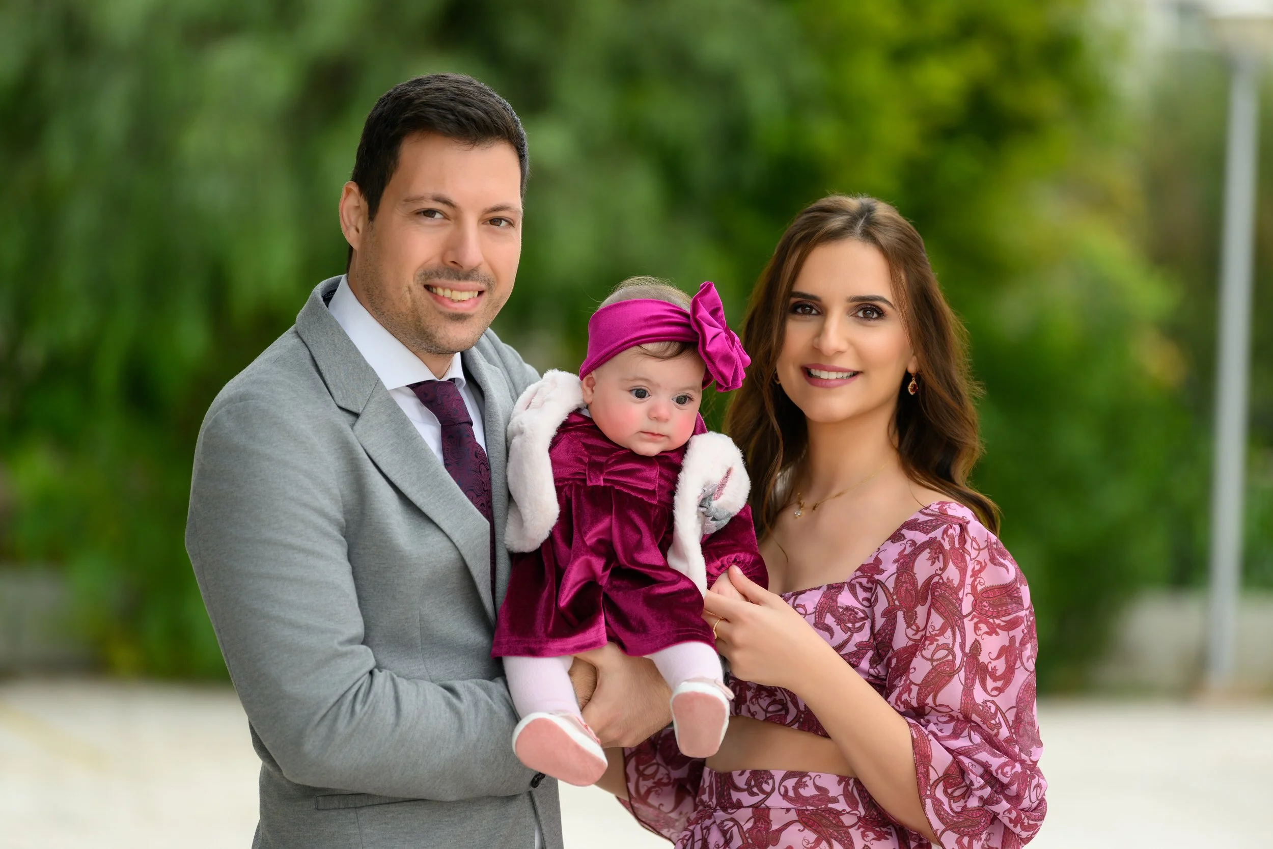 A family of three, a man, a woman, and a baby girl, standing outdoors in a park with green foliage in the background. The man is wearing a gray suit and white shirt, the woman is in a pink patterned dress, and the baby girl is dressed in a pink velvet dress with a matching headband, being held by the man.