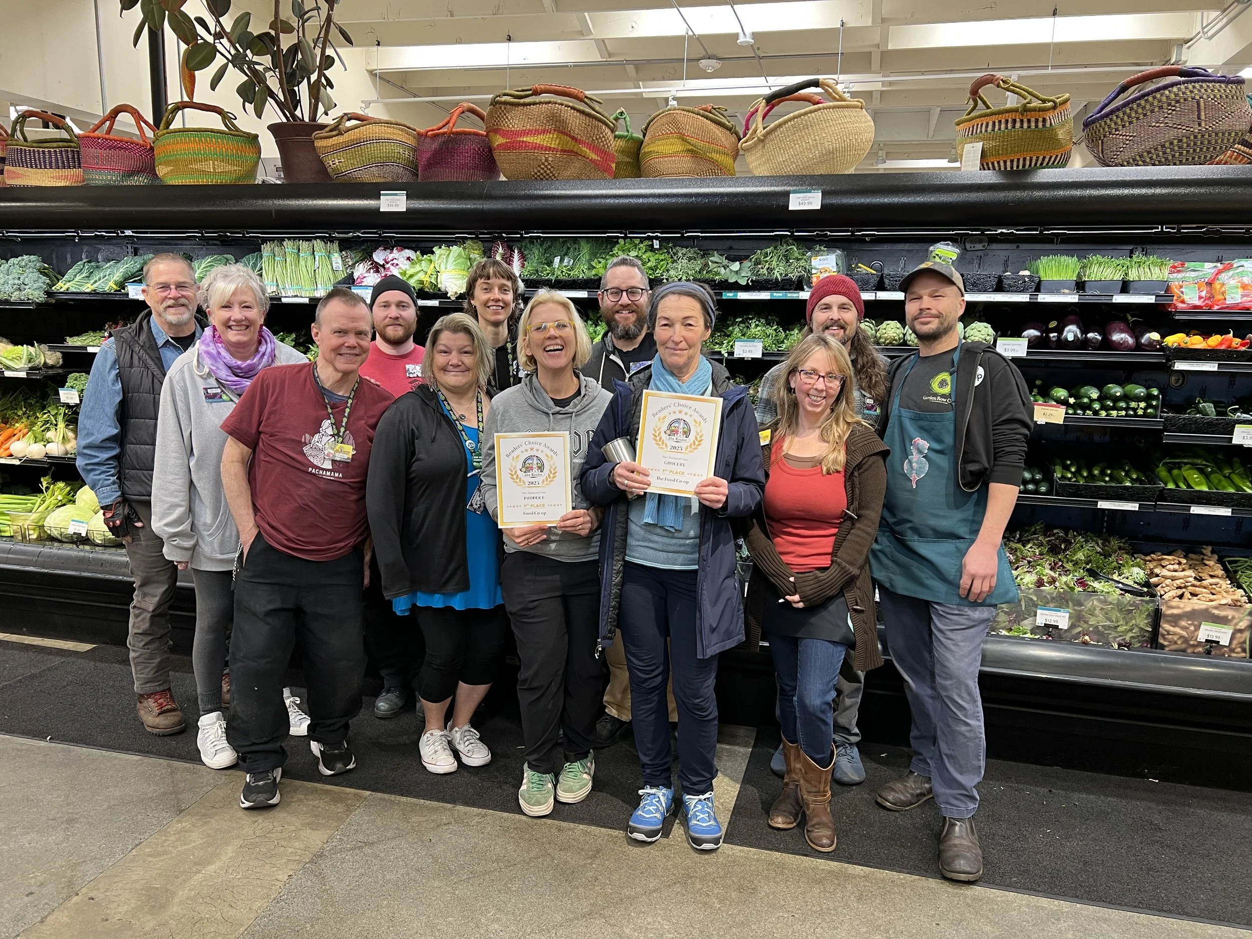 A group of twelve people standing together in a grocery store aisle, smiling and holding awards, with fruits and vegetables displayed on shelves behind them.