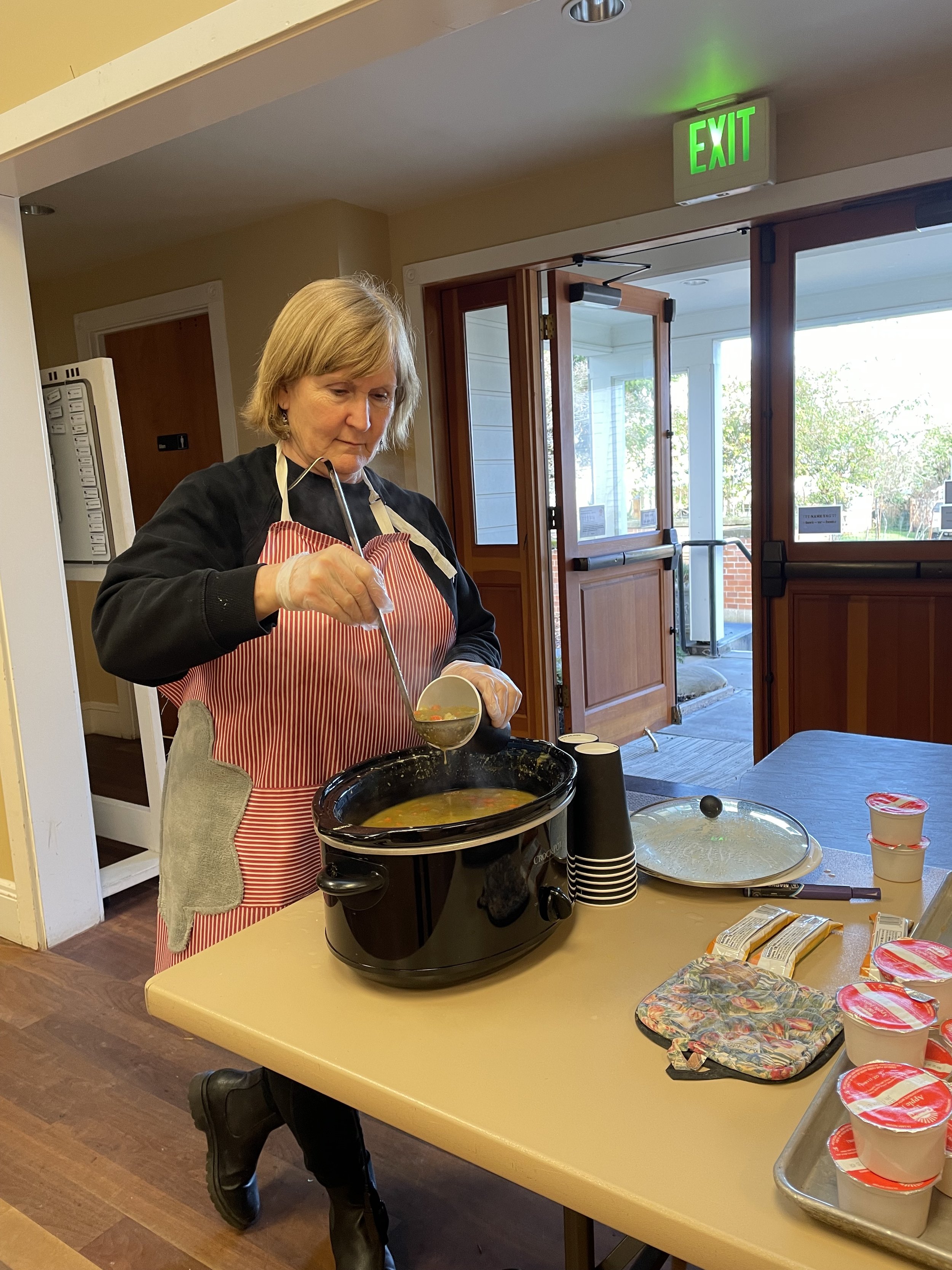 Woman serving soup from a slow cooker at a table, with yogurt cups nearby, inside a room with open door and a visible exit sign.