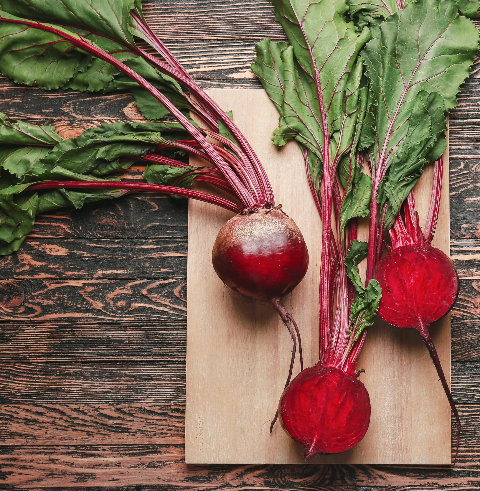 Three beets with green leaves on a wooden cutting board and rustic wooden table.