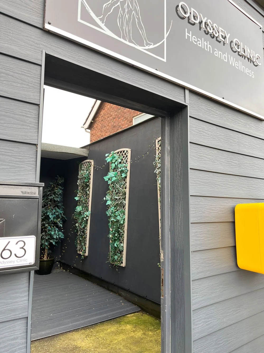 Exterior view of the Odyssey Clinic for health and wellness, with clinic's sign on the front, number 63, plants and a grey door.
