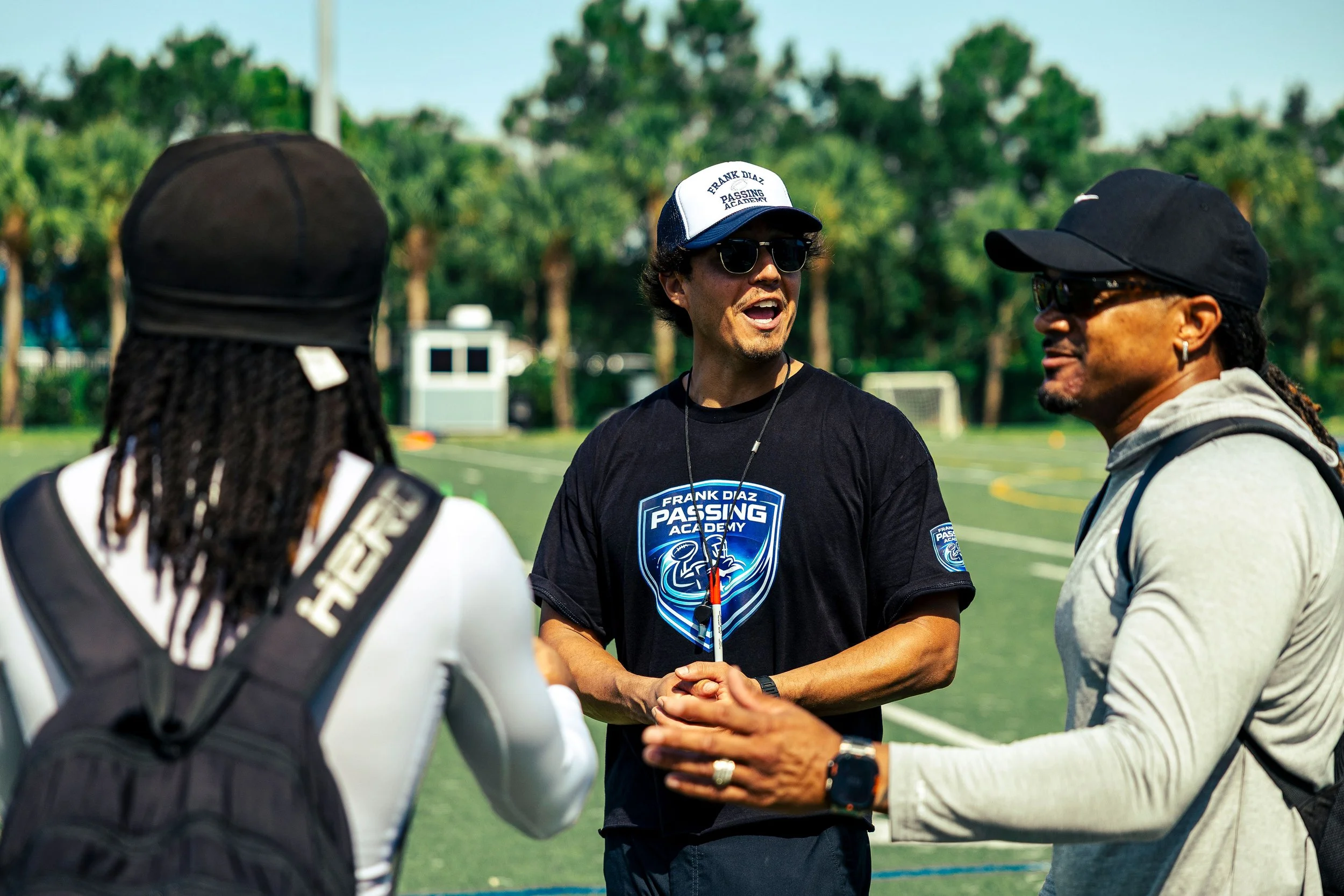 Three men standing in a circle outdoors on a sports field, engaged in conversation. One man is wearing sunglasses, a cap that says 'Frank Diaz Passing Academy,' and a black t-shirt with a logo. The other two men are wearing caps and casual clothing. In the background, there are trees and sports equipment.