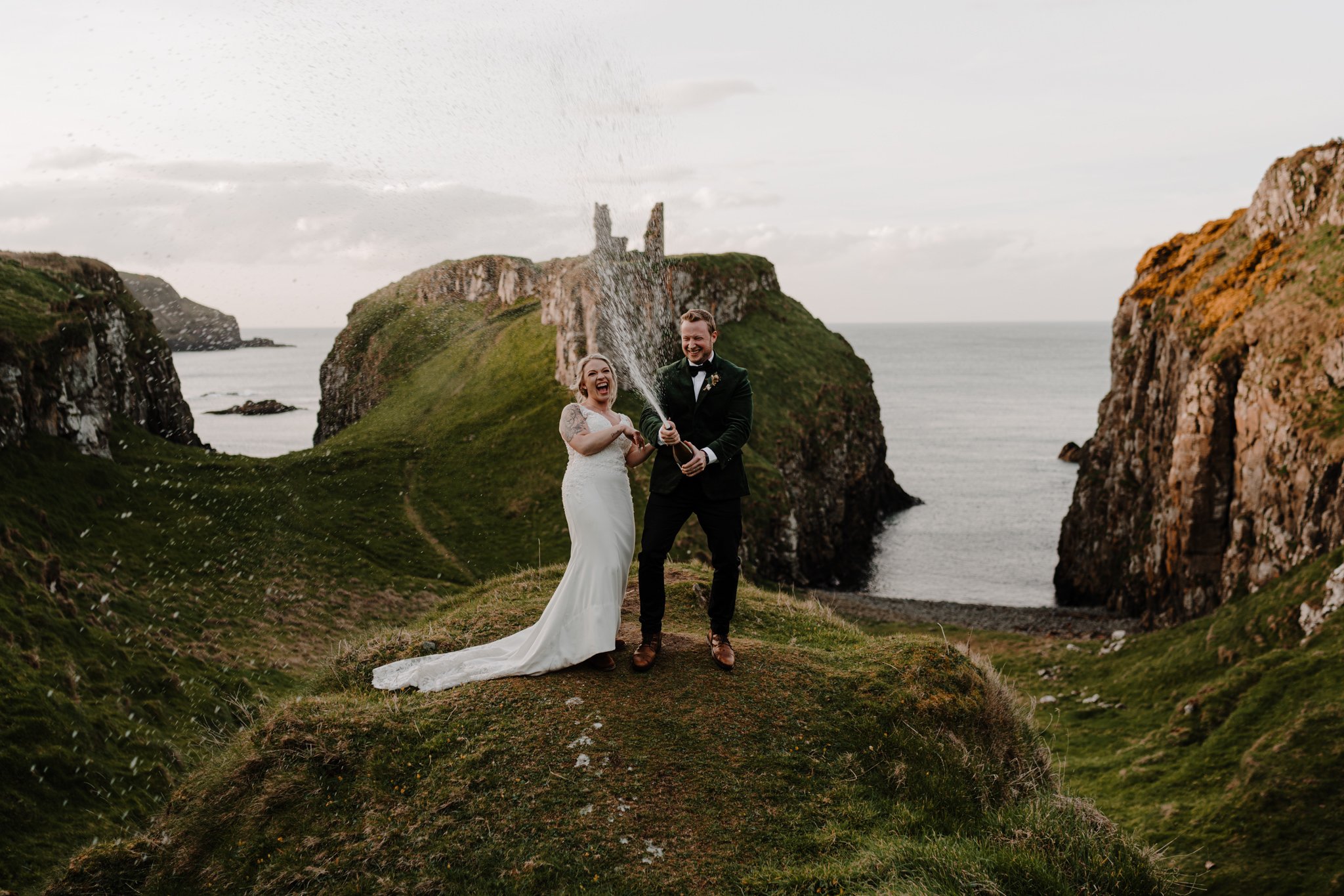 Bride and groom celebrating with champagne on a grassy cliff overlooking the ocean, with green hills and rocky formations in the background, during their wedding.