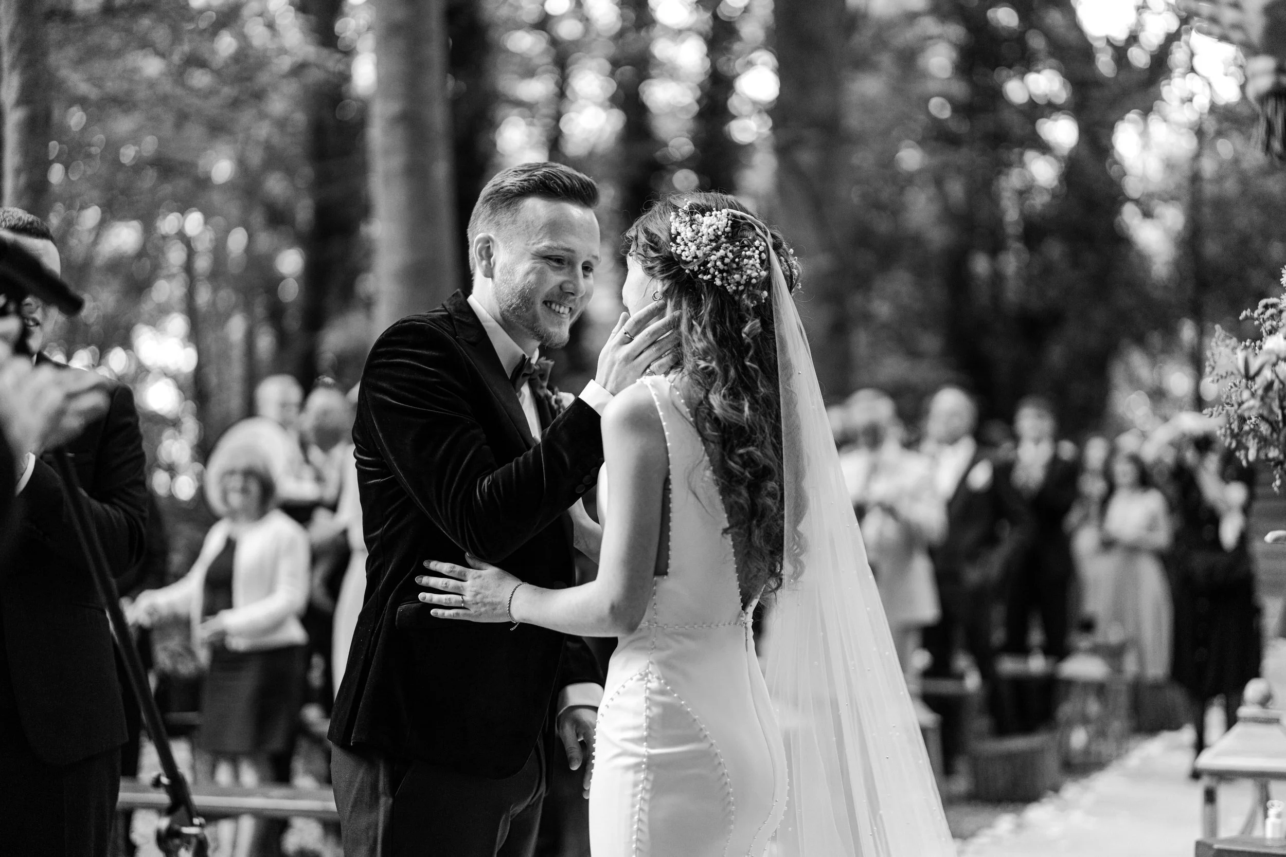 A black-and-white photograph captures a bride and groom sharing a kiss during their wedding ceremony outdoors, with guests in the background.