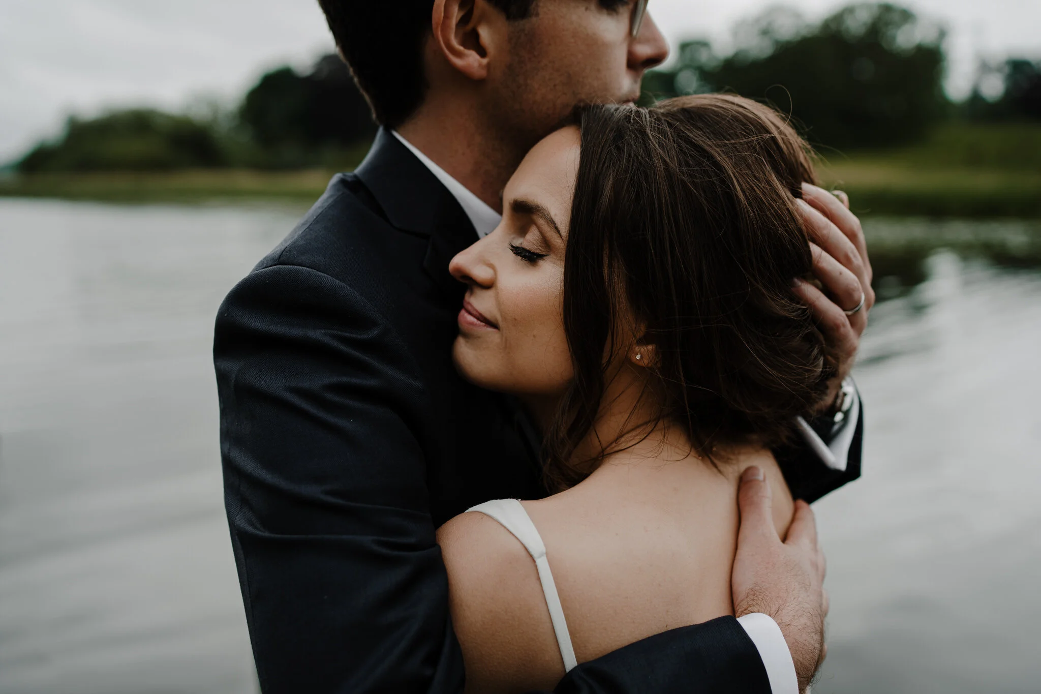 A man in a suit gently embracing a woman near a body of water during daytime. The woman has her eyes closed and is smiling slightly, with her head resting on the man's chest. They are outdoors with greenery in the background.